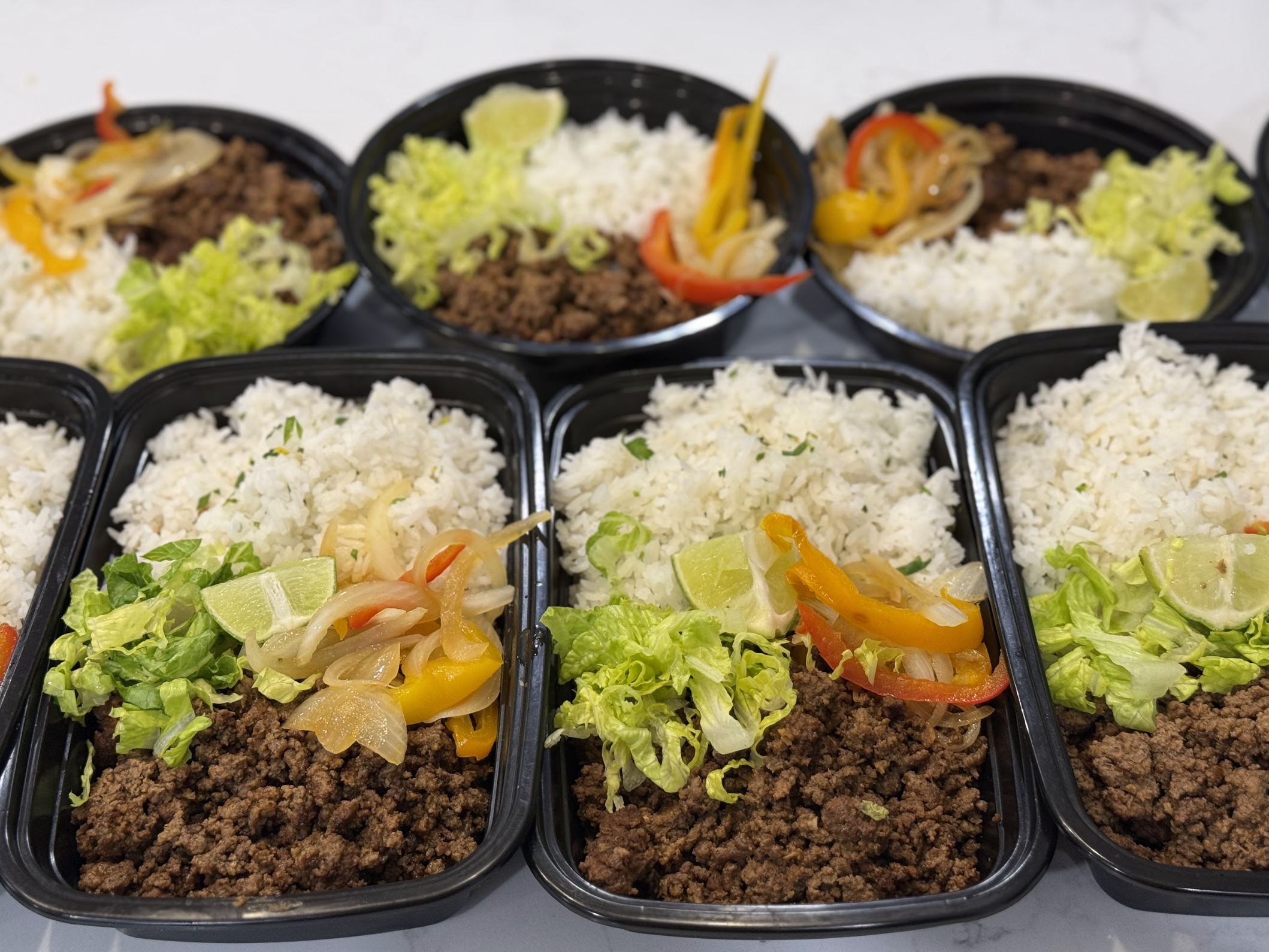Meal prep containers with cooked ground beef, rice, and vegetables like lettuce, bell peppers, onions, and lime wedges.