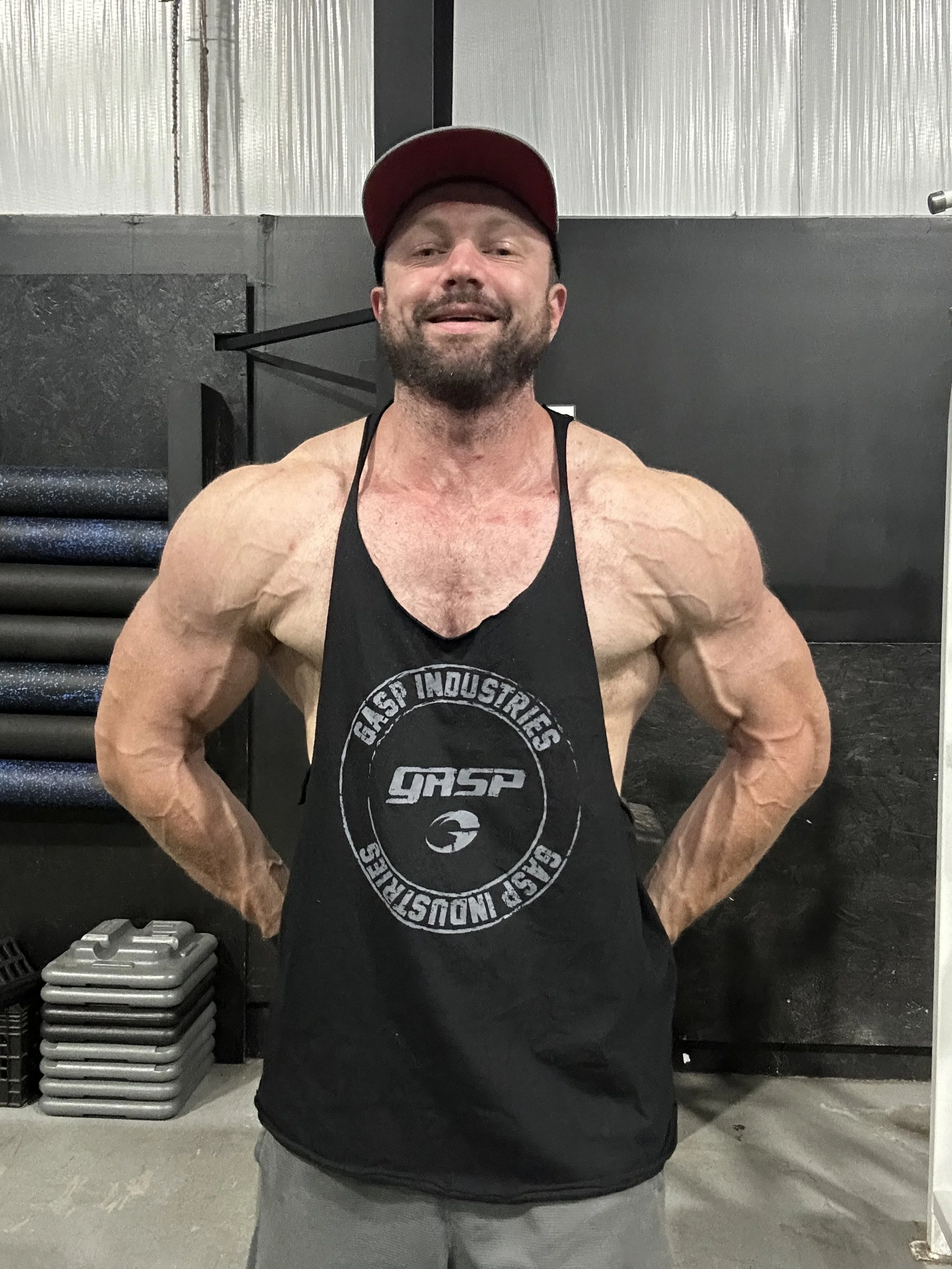 A muscular man with a beard wearing a black tank top and a red cap, smiling at the camera in a gym.