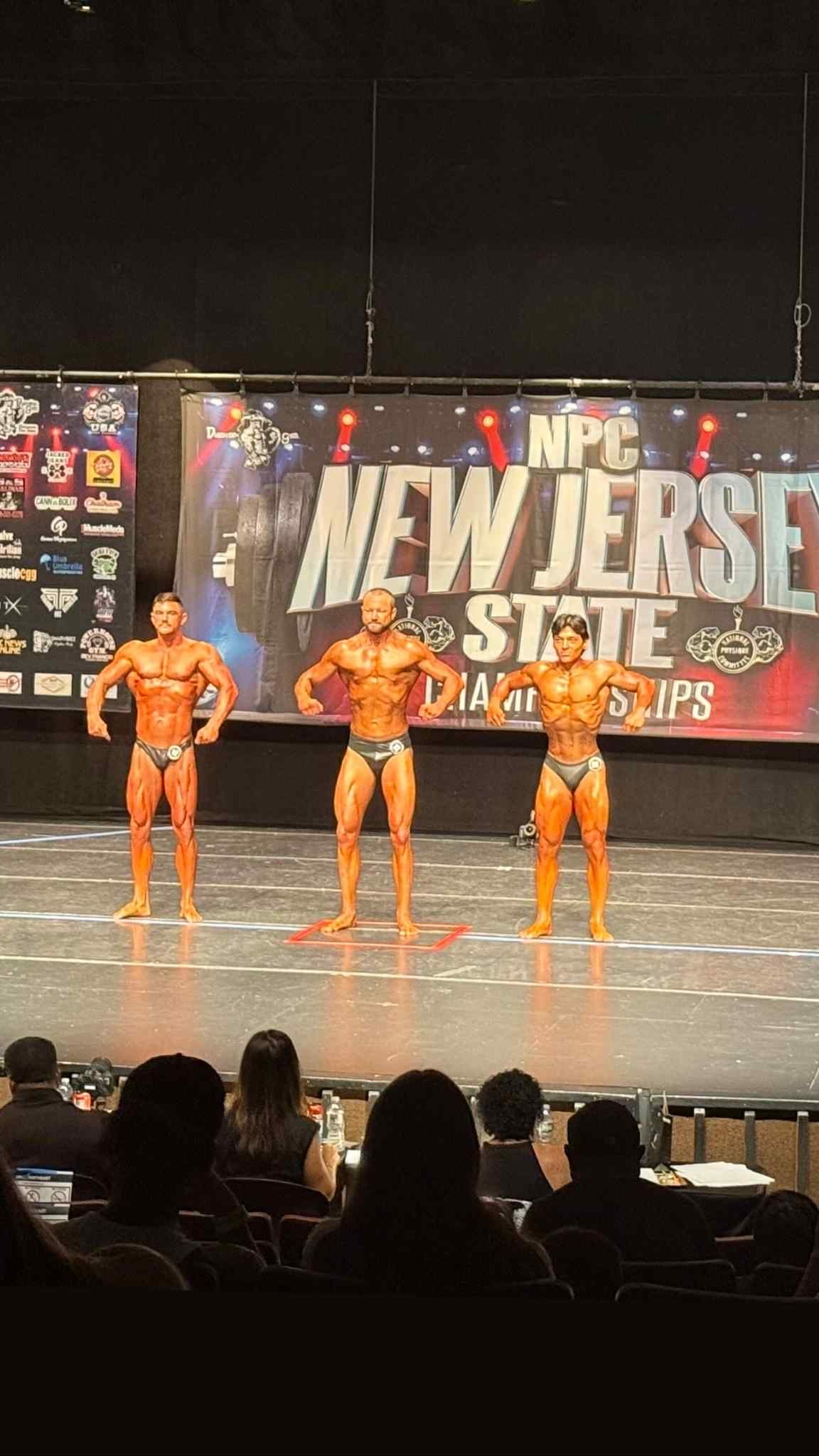 Three male bodybuilders in posing trunks compete on stage at the NPC New Jersey State Championships, standing in front of a large banner with the competition's name.