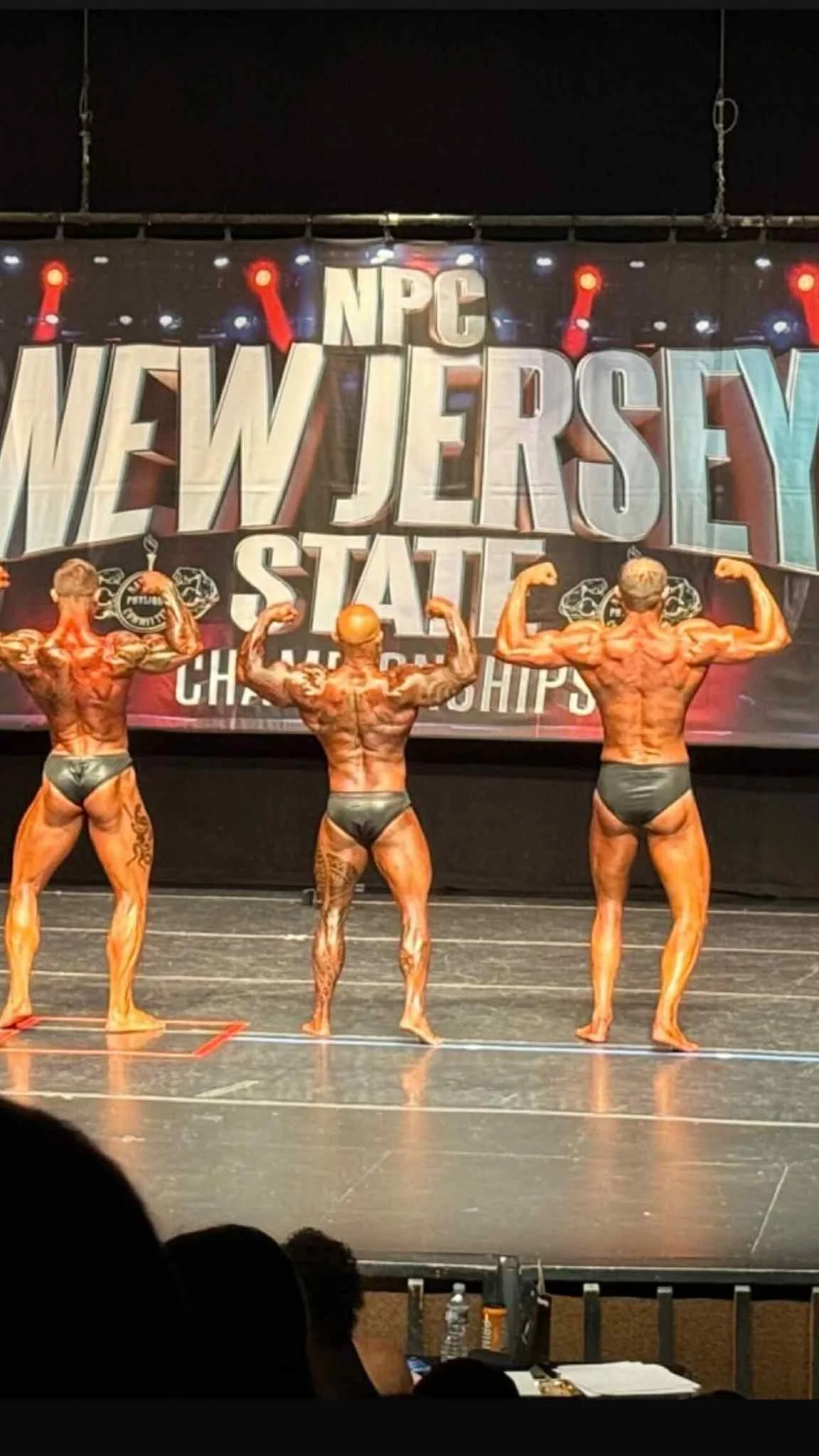 Three male bodybuilders on stage, flexing their muscles during a bodybuilding competition at the NPC New Jersey State Championships.