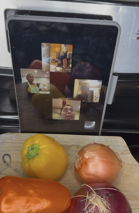 A tablet mounted on a black surface displaying a video call with four people, and a wooden cutting board with yellow bell pepper, onion, red onion, and orange bell pepper in front of it.