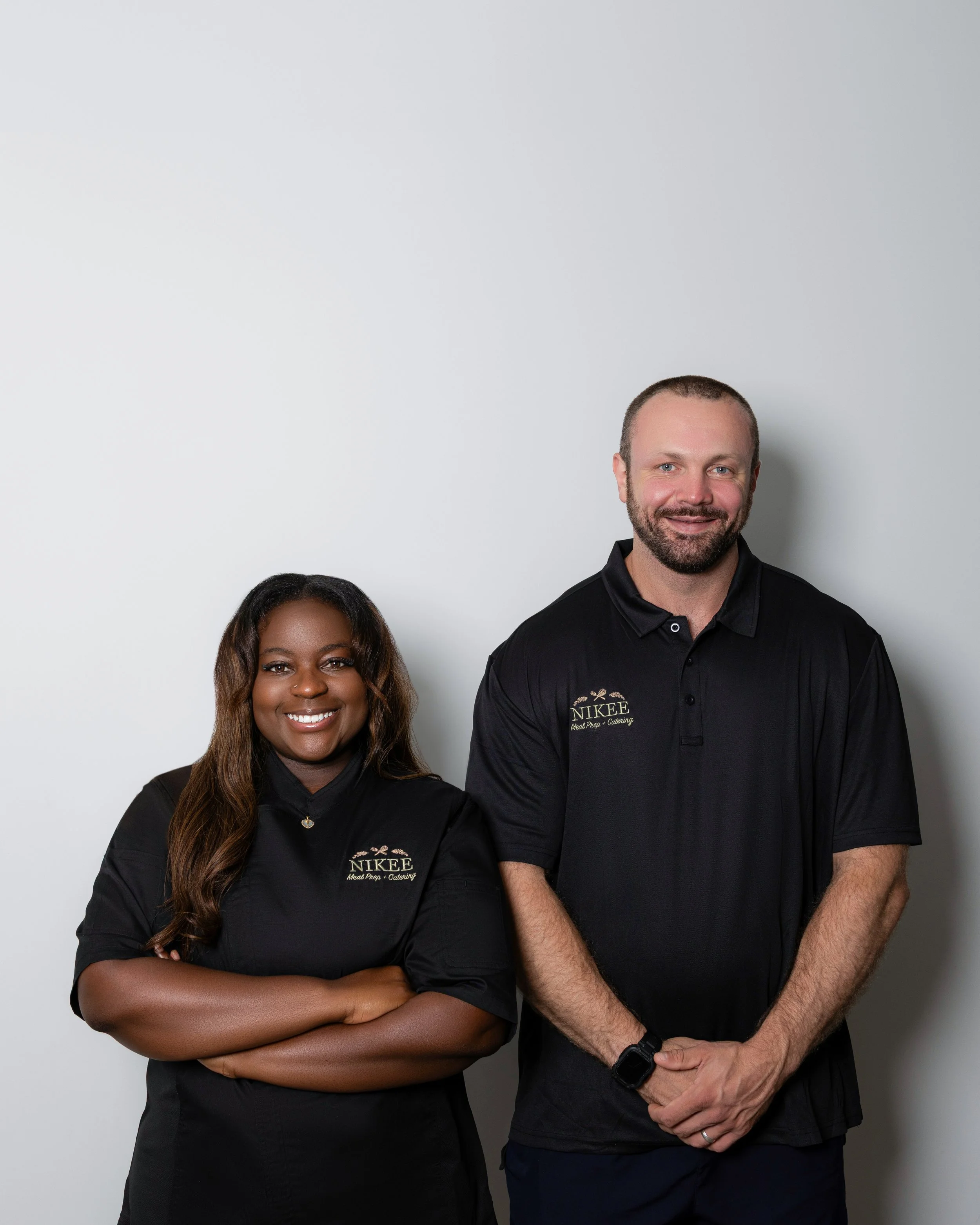 Two people standing against a plain white wall. The woman is on the left with arms crossed, smiling, wearing a black shirt with a logo on it. The man is on the right with hands clasped in front, smiling, wearing a black polo shirt with a logo on it.