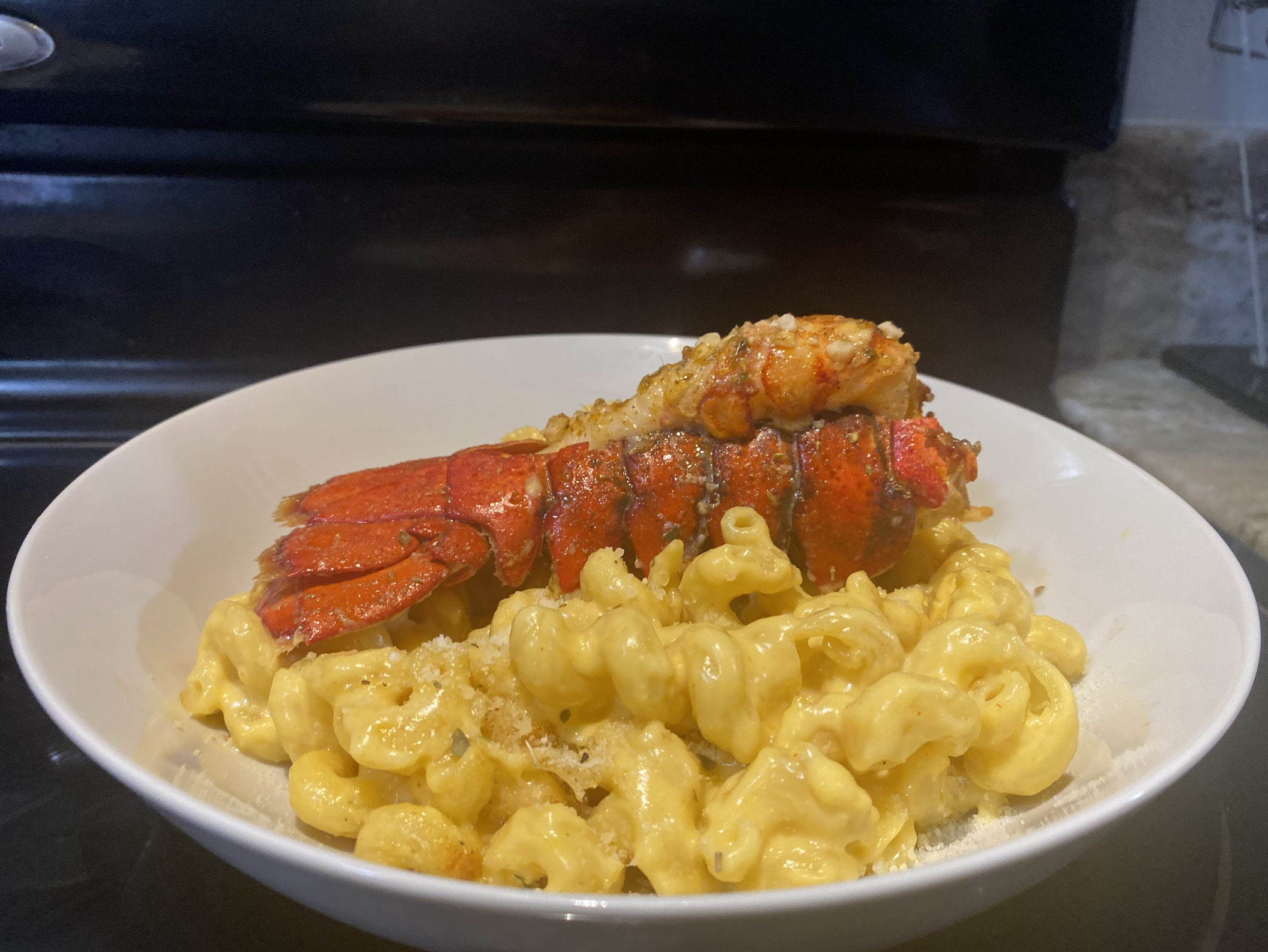 A bowl of creamy macaroni and cheese topped with lobster tail and herbs, with part of a black kitchen appliance in the background; a dish by Nikee Catering.