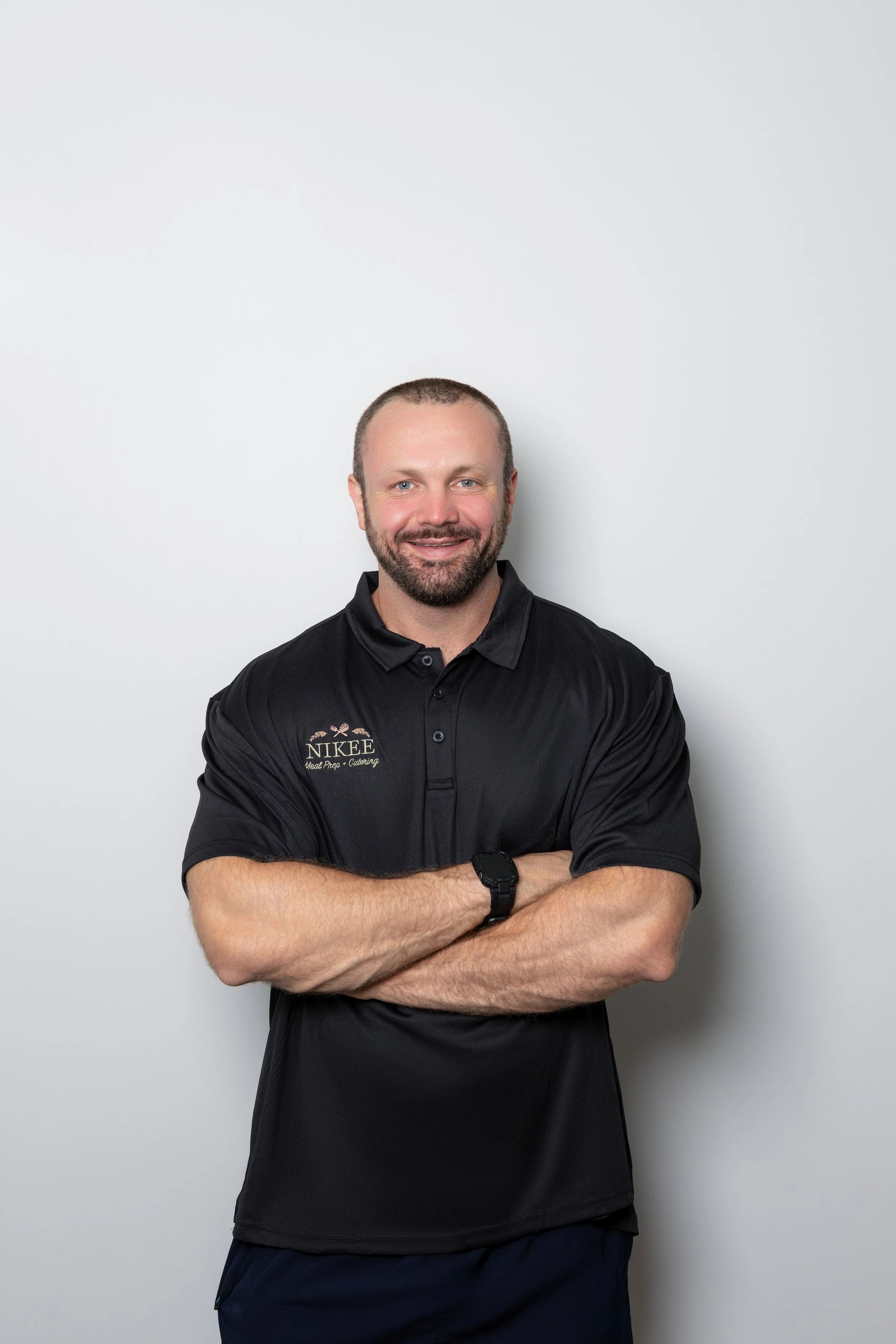 A smiling man with a beard and short hair, wearing a black polo shirt with a Nikee Meal Prep & Catering logo and dark shorts, standing with arms crossed against a plain gray background.