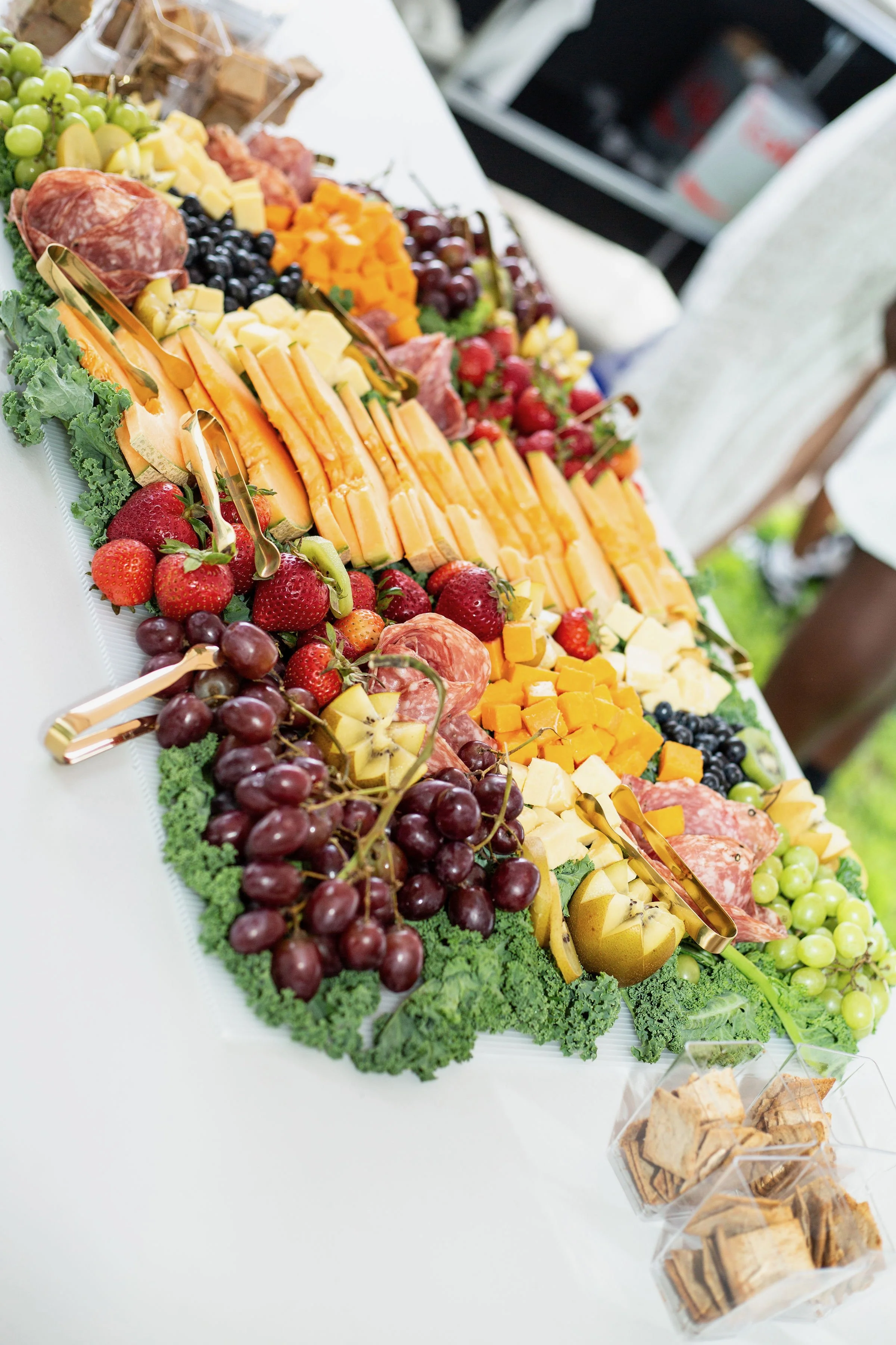 A large platter of assorted fruit, cheese, and cured meats arranged with grapes, strawberries, diced melon, and leafy greens; gold tongs are placed on the platter for serving. Two containers of crackers are visible in the lower right corner.