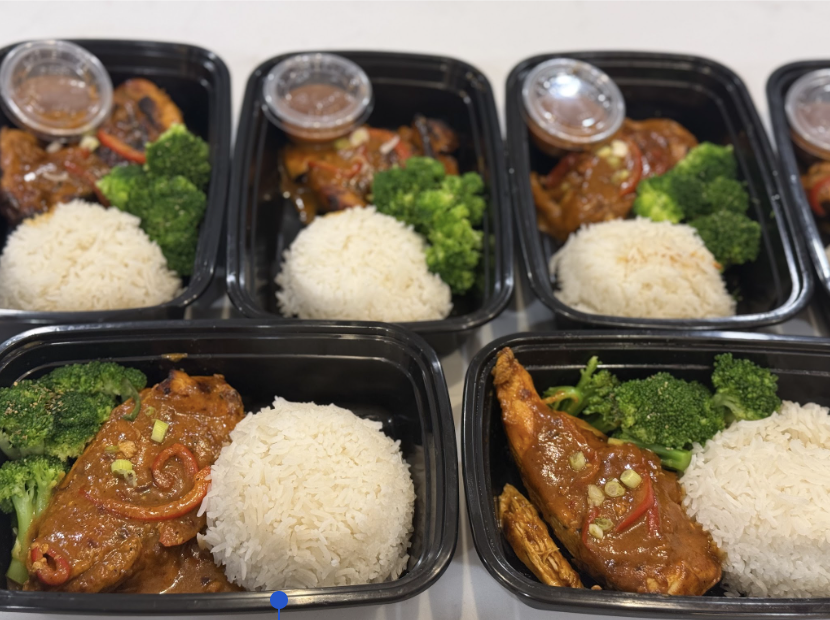 Pre-packaged black containers with rice, broccoli, and glazed chicken drumsticks topped with green onions, accompanied by small containers of sauce.
