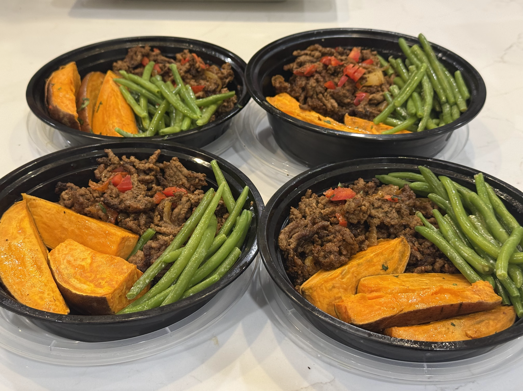 Four black takeout bowls filled with cooked ground beef, green beans, and roasted pumpkin slices, placed on a white table.