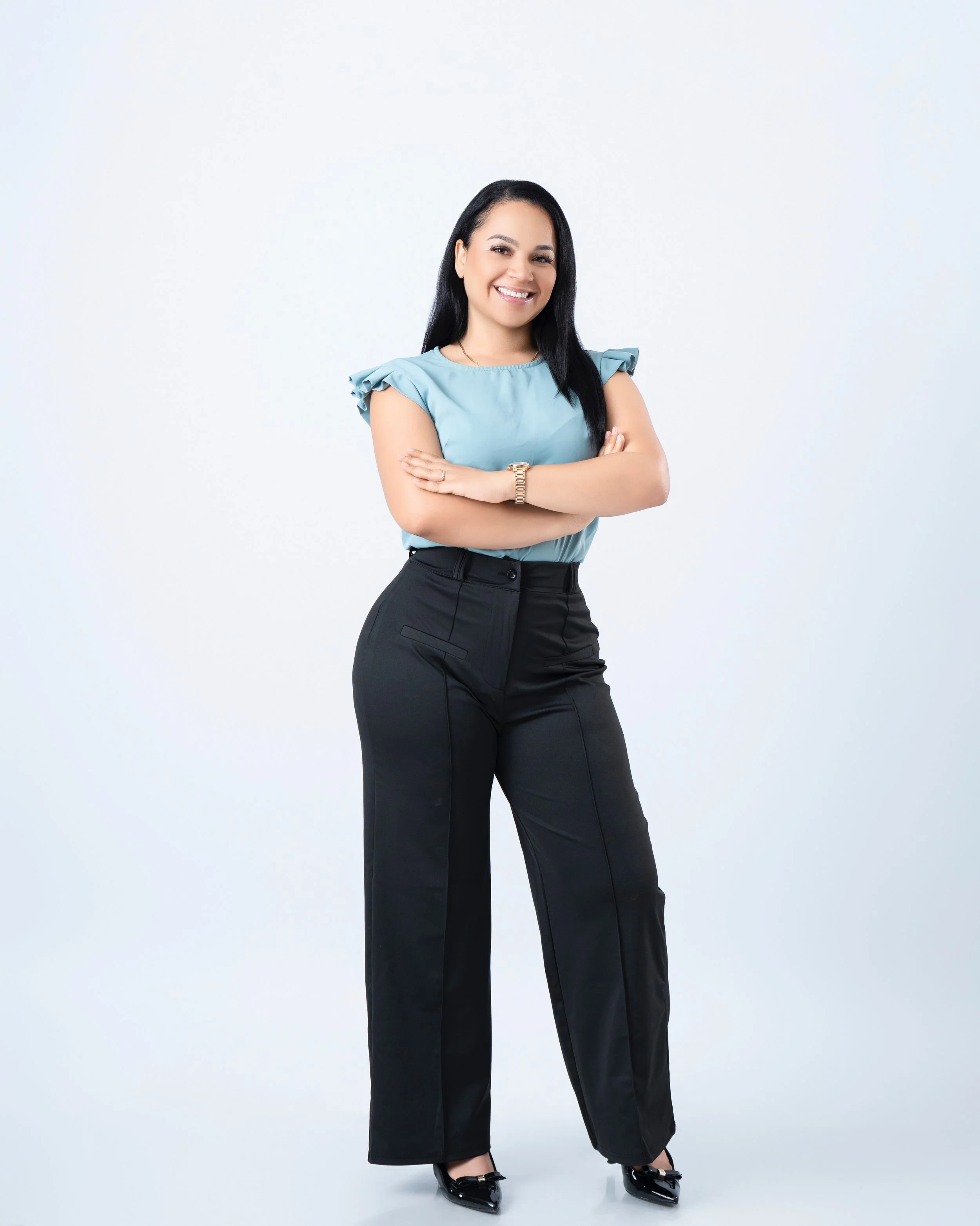 A woman with black hair, wearing a blue top and black pants, standing with her arms crossed and smiling at the camera.