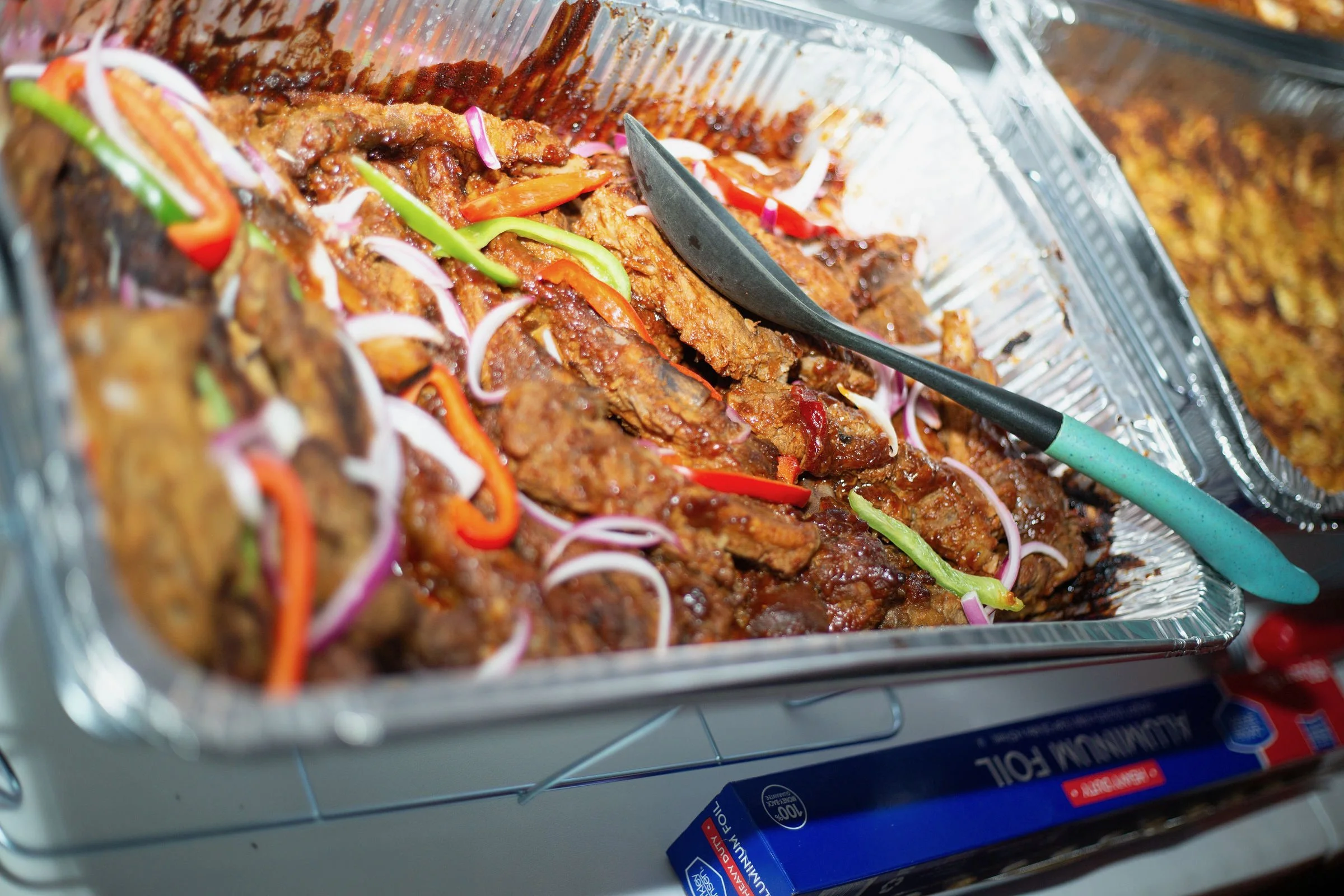 A close-up of cooked meat with barbecue sauce, garnished with sliced red onions, green onions, and red bell peppers, inside a foil container on a portable stove or grill, catered by Nikee Catering.