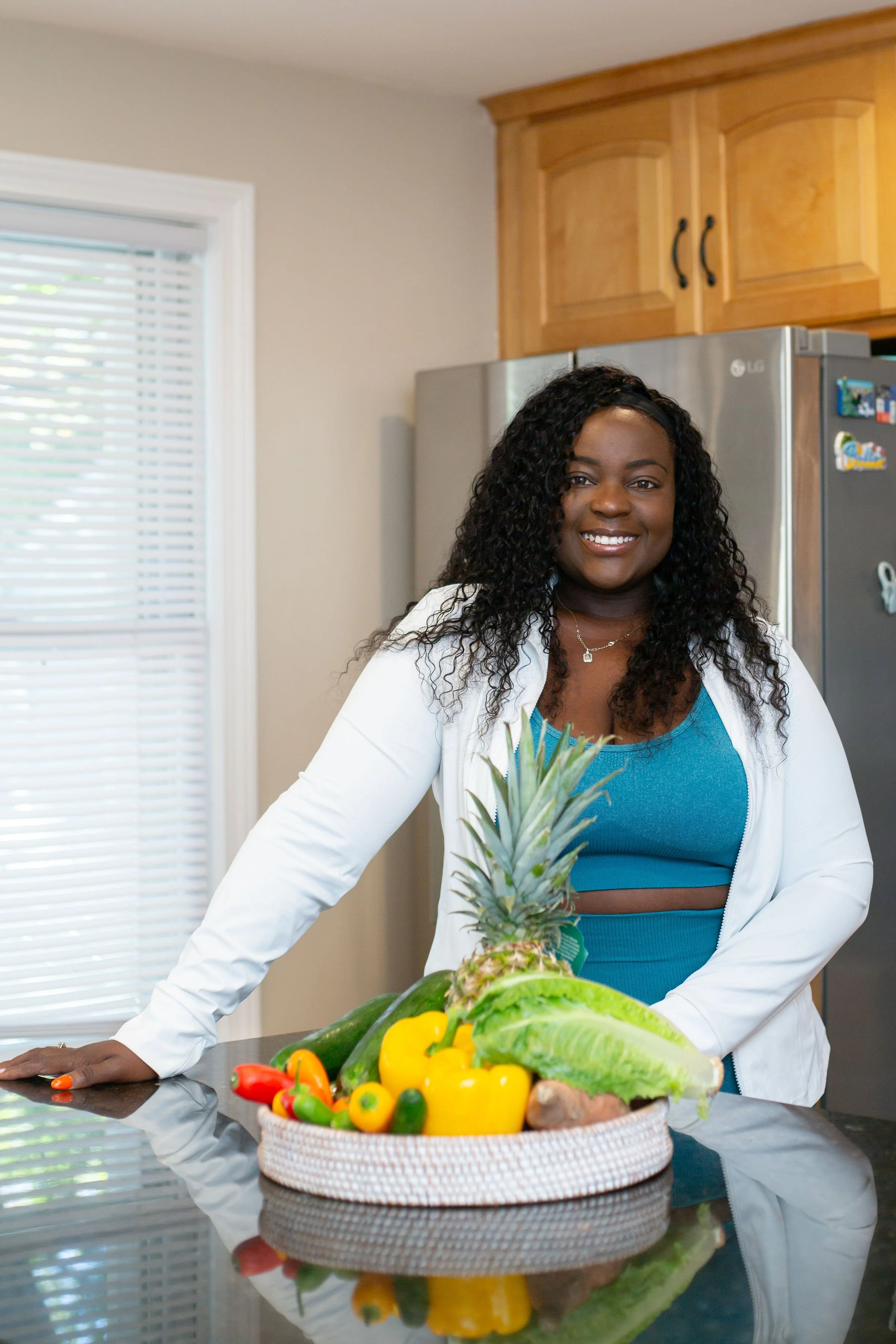 A woman in a white jacket standing in a kitchen with a basket of fresh fruits and vegetables, including pineapple, bell peppers, cucumbers, lettuce, and ginger, on a reflective countertop.