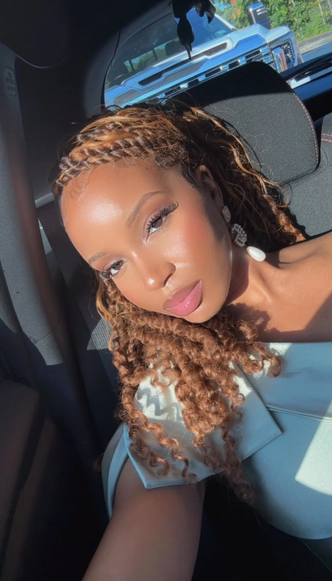 A woman with curly, reddish-brown hair taking a selfie inside a car, with sunlight illuminating her face.