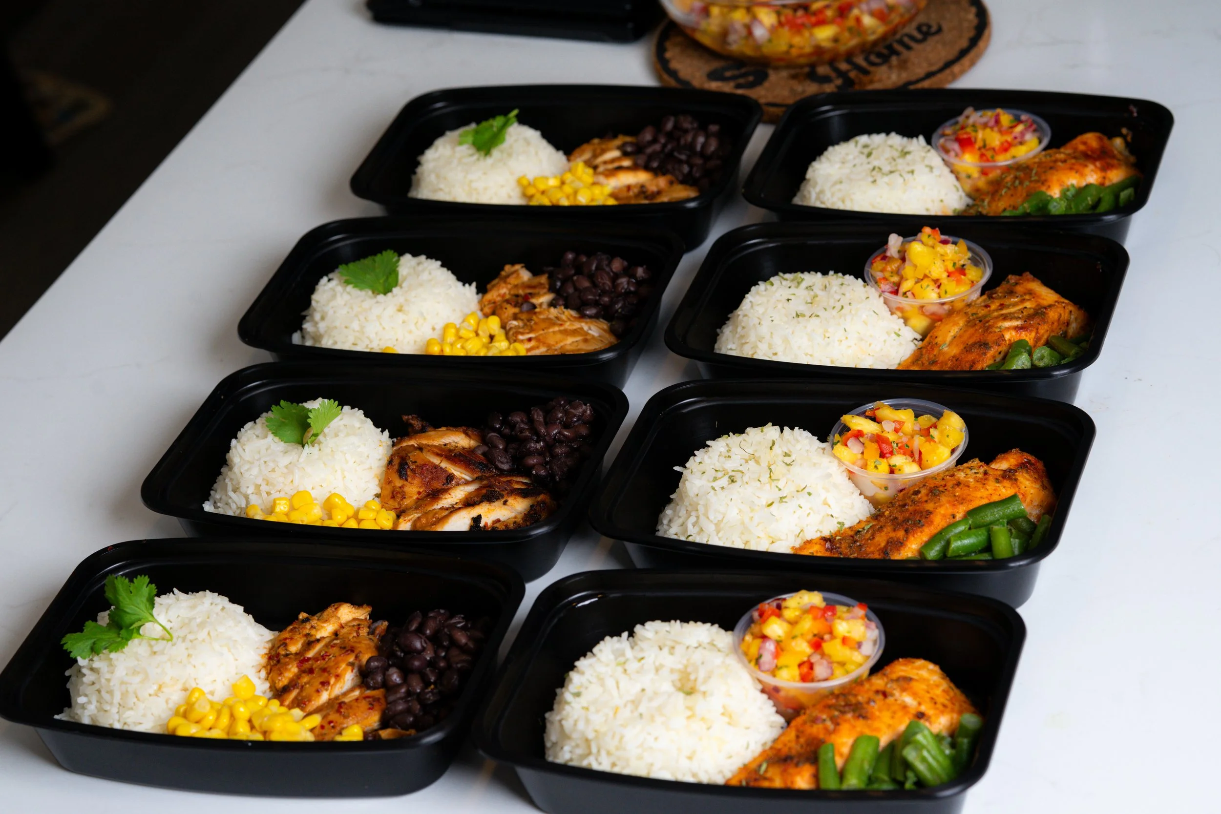 Multiple black meal prep containers filled with white rice garnished with cilantro, black beans, grilled chicken, yellow corn, a small cup of colorful Mexican corn salad, and cooked green beans on a white surface.