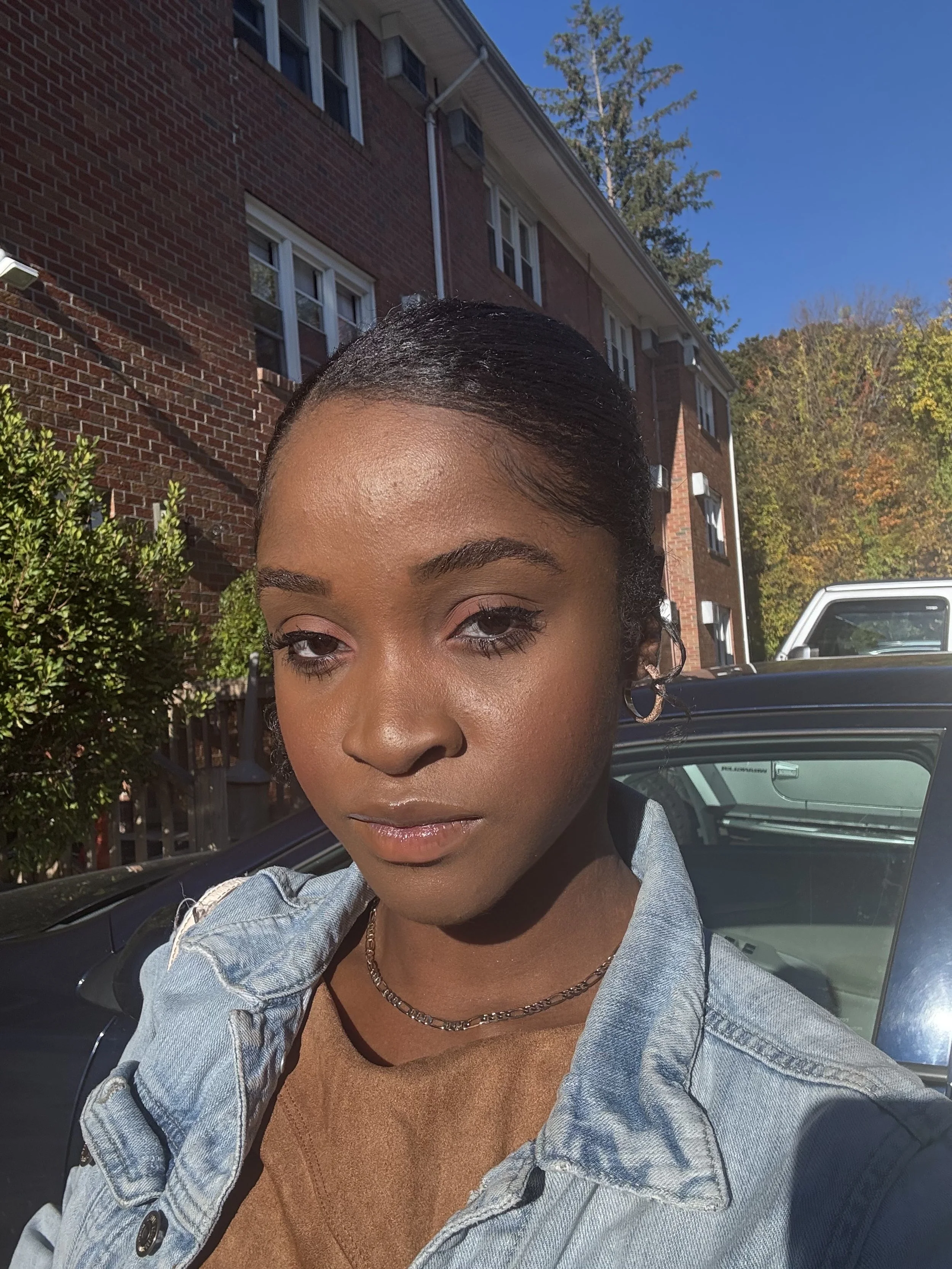 A young woman taking a selfie outside on a sunny day, with a brick apartment building and trees with fall foliage in the background.
