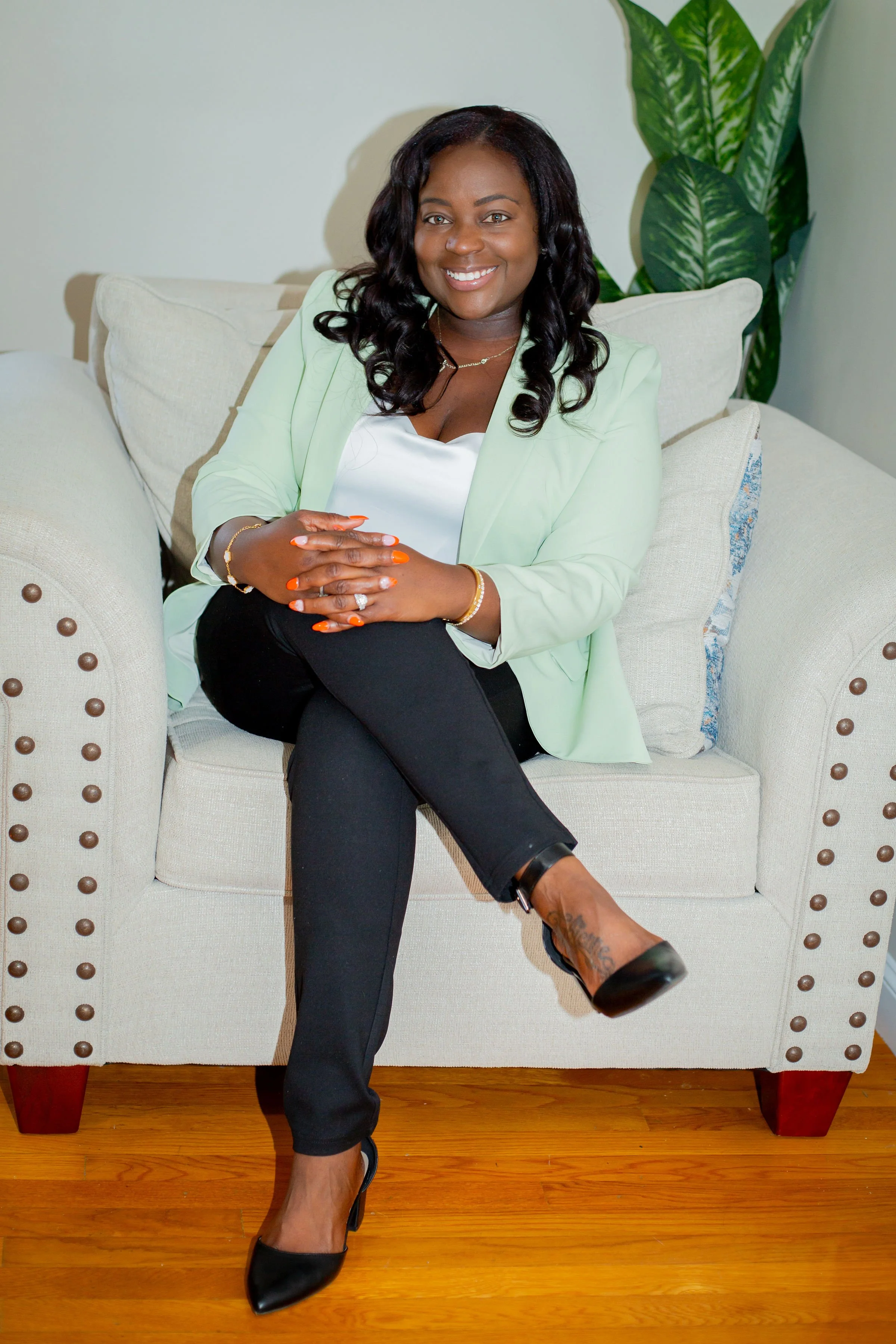 A woman with dark wavy hair sitting on a beige sofa with multiple pillows, smiling and looking at the camera. She is wearing a light green blazer, white top, black pants, and black high heels. She has tattooed ankles and is accessorized with gold jewelry and orange nail polish. A green plant is in the background.