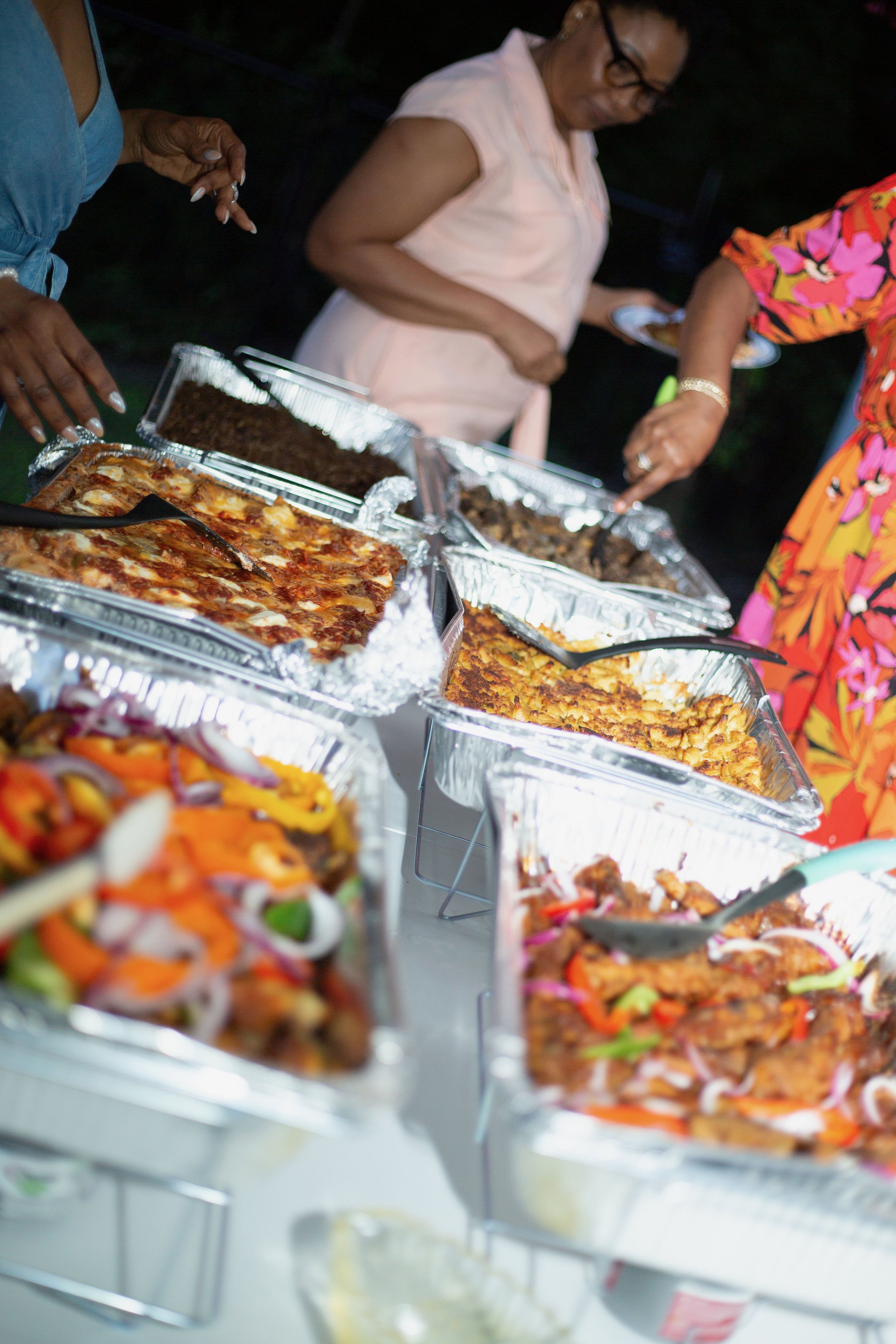 Multiple aluminum trays filled with various cooked dishes such as pizza, pasta, and meat, with people serving themselves at a buffet table outdoors, catered by Nikee Catering.