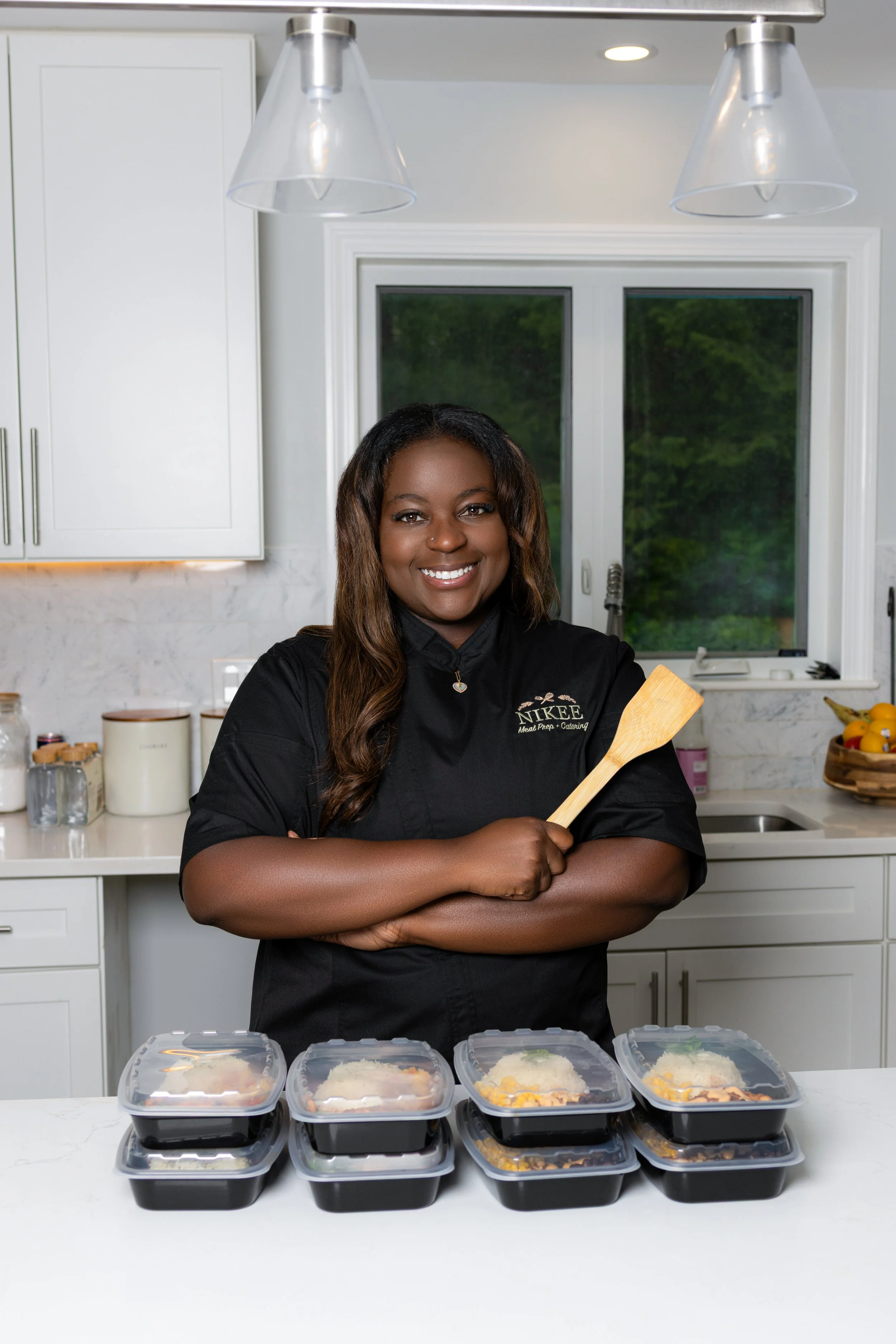 A woman standing in a kitchen, smiling and holding a spatula, wearing a shirt with a Nikee Meal Prep & Catering logo on it, with multiple meal prep containers on a white countertop in front of her.