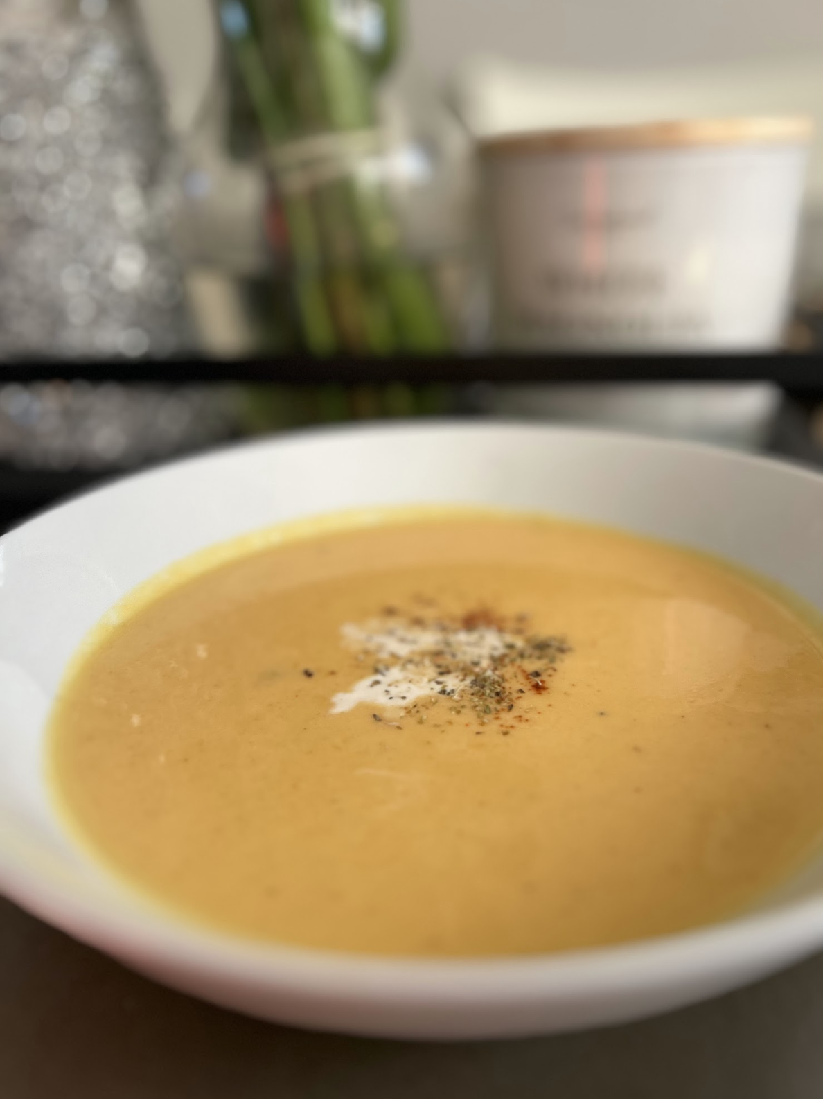 Creamy orange soup in a white bowl, topped with herbs and spices, with a blurred kitchen counter and flower vase in the background; a dish by Nikee Catering.
