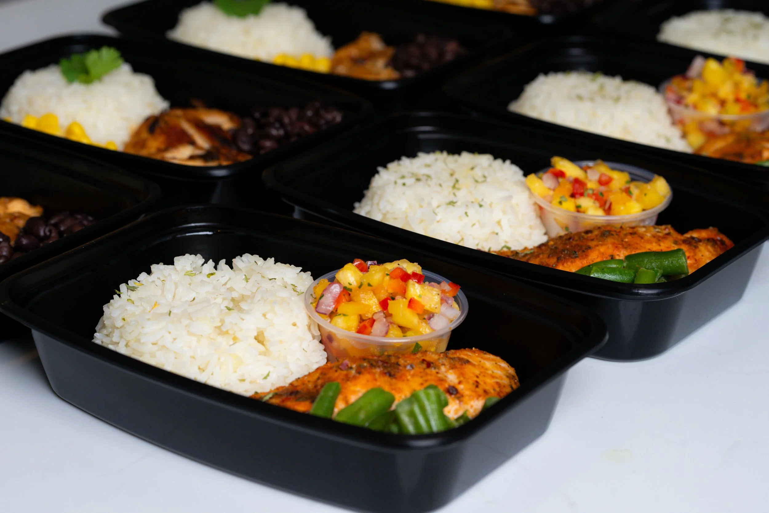 Multiple black meal prep containers filled with white rice, cooked chicken, black beans, and a mixed vegetable salad with yellow, red, and green peppers, served with lime wedges.