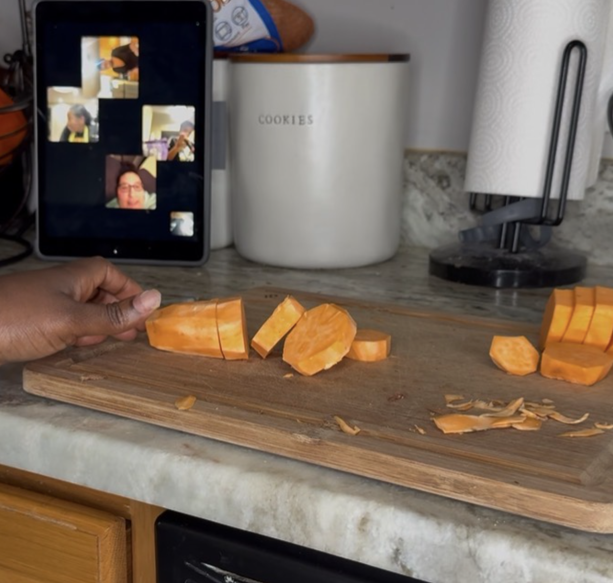 A person's hand holding a sliced orange pumpkin on a wooden cutting board, with additional pumpkin slices and peels, in a kitchen setting. In the background, an iPad showing a video call, a white jar labeled 'COOKIES,' a paper towel roll, and kitchen utensils.