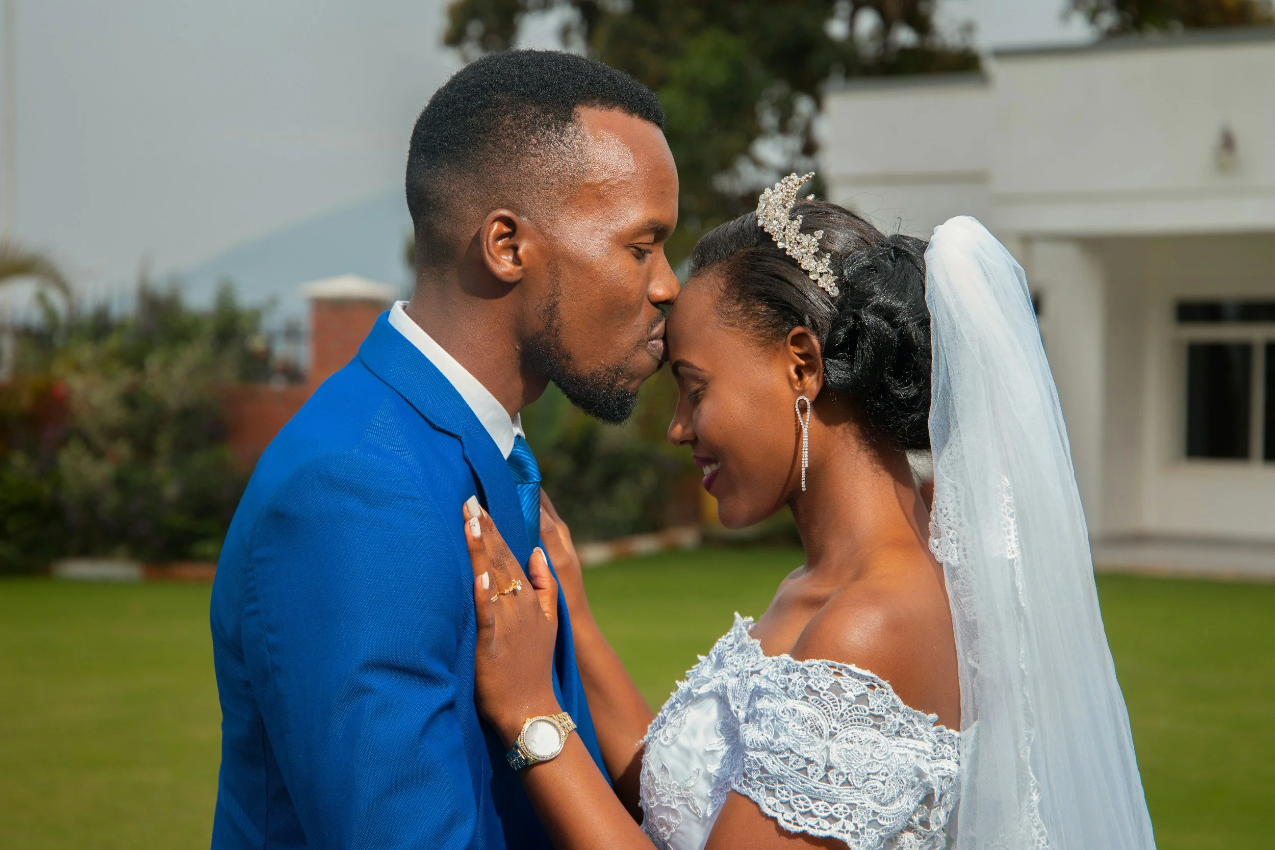A bride and groom touching foreheads outdoors during wedding, dressed in traditional wedding attire with the groom in a blue suit and the bride in a white lace dress with veil and tiara.