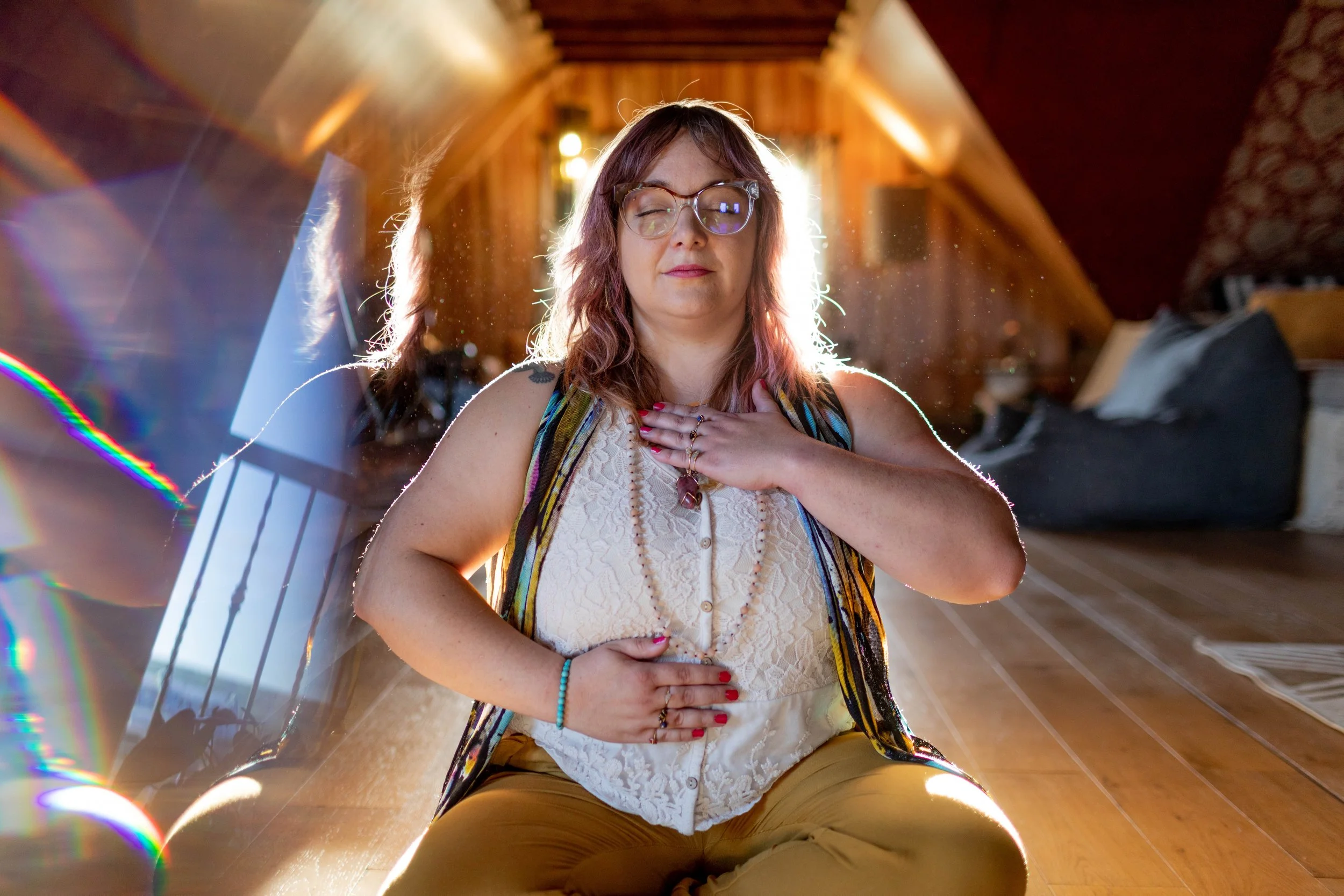 A woman with glasses and shoulder-length wavy hair, sitting cross-legged on a wooden floor with sunlight streaming behind her. She has her eyes closed, one hand on her chest, and the other on her stomach, appearing to meditate or practice yoga. The room has wooden walls and a warm, cozy atmosphere.