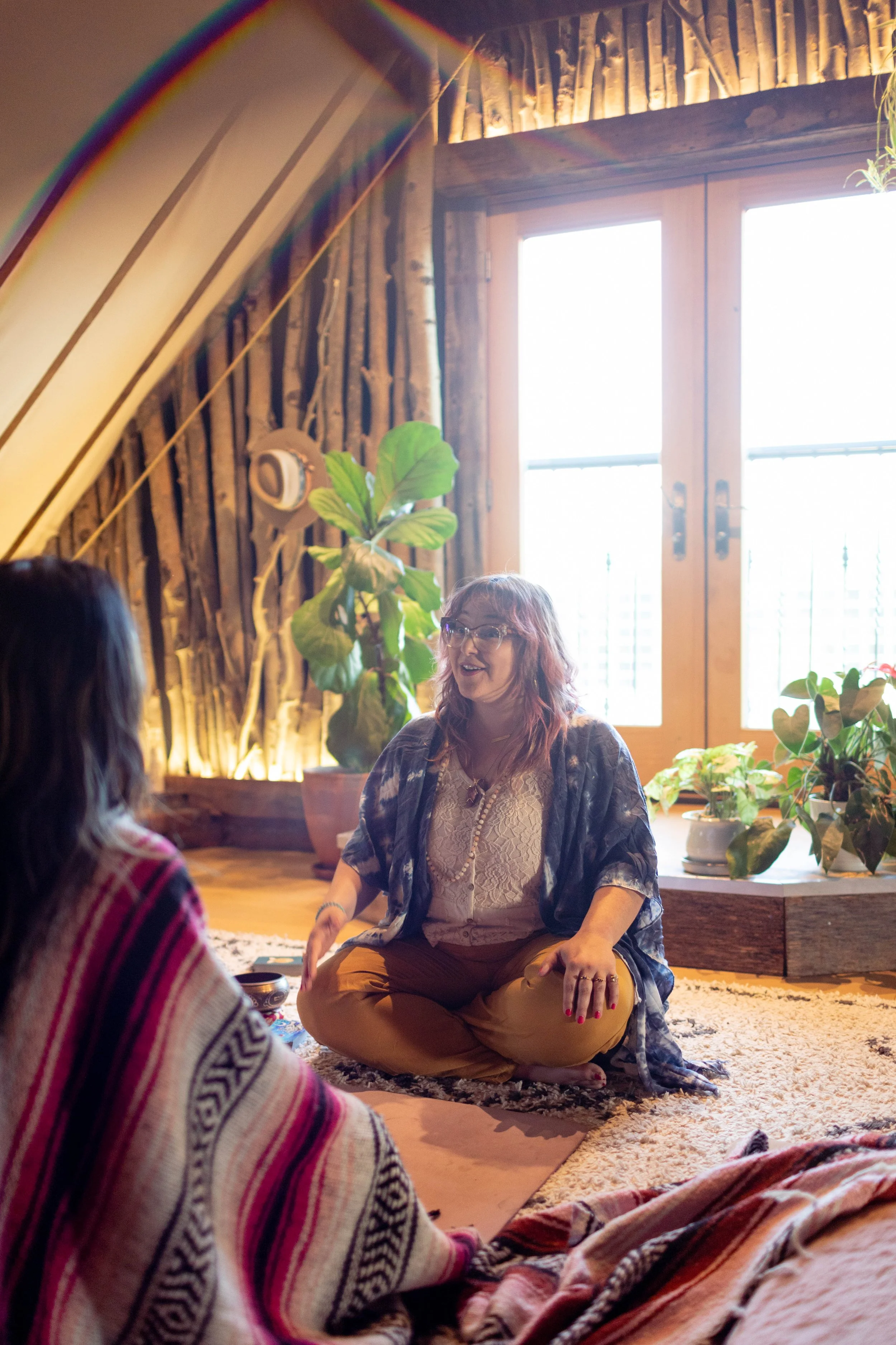 A woman with glasses and reddish hair sitting cross-legged on a carpeted floor, engaging in conversation with a person in a patterned sweater. The room has large windows, wooden decor, and various green plants.