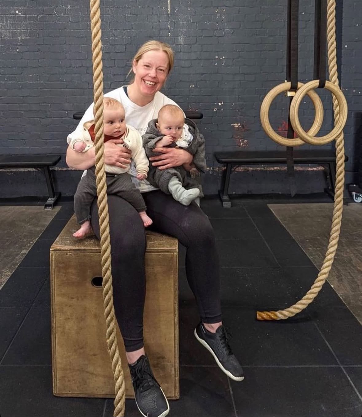 A woman sitting on a wooden box, holding two babies in baby carriers, inside a gym with gymnastics rings and a rope in the background.