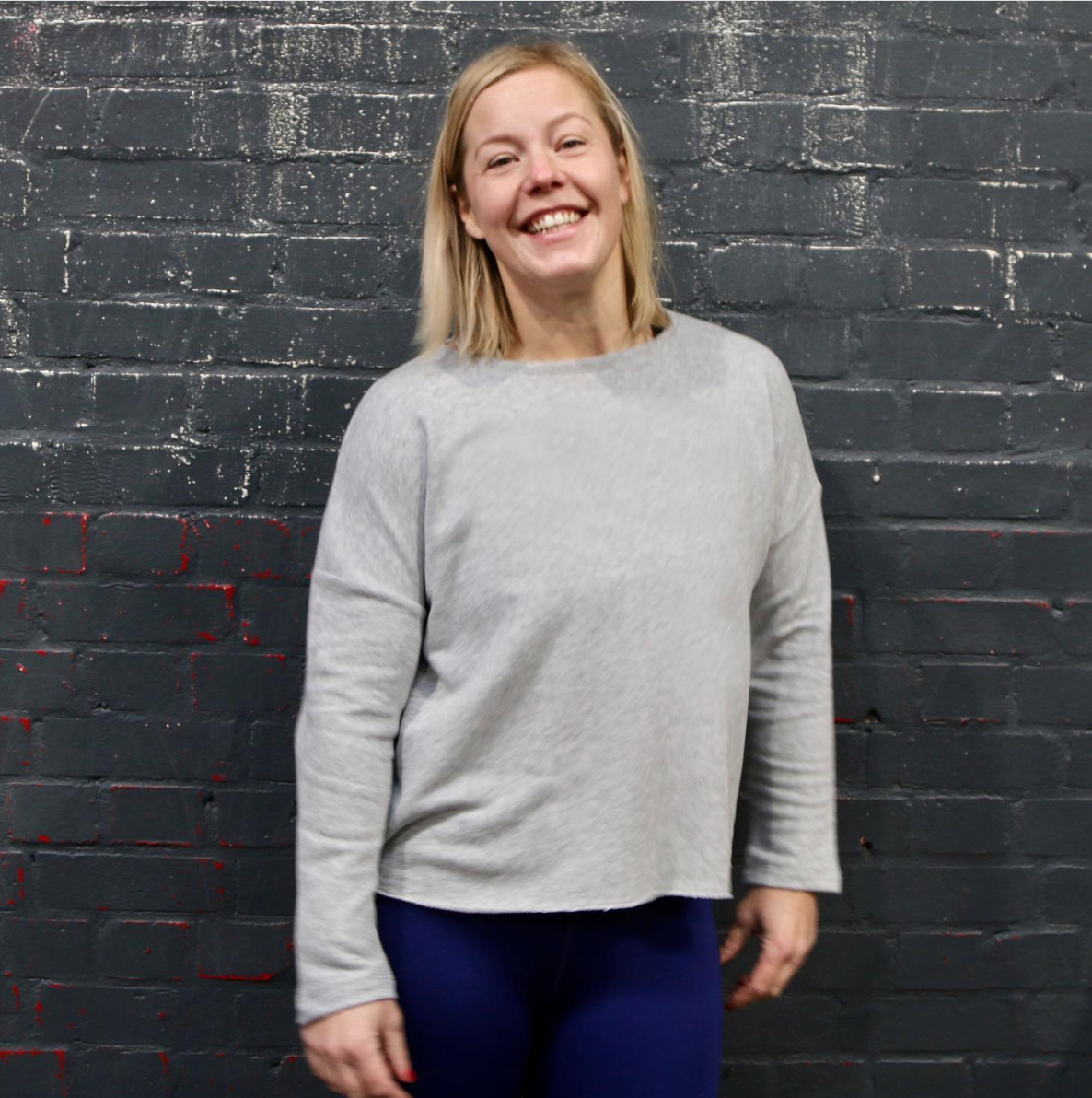 A smiling woman with shoulder-length blonde hair standing against a dark gray brick wall, wearing a light gray sweater and dark pants.