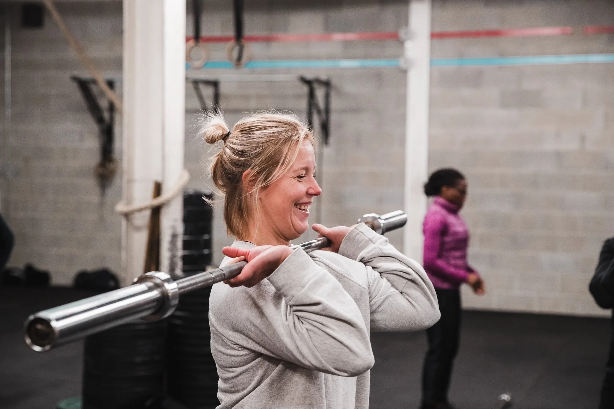 A woman with blonde hair tied in a small bun, smiling while holding a barbell across her shoulders during a workout in a gym. In the background, a person in a pink jacket and glasses is visible.