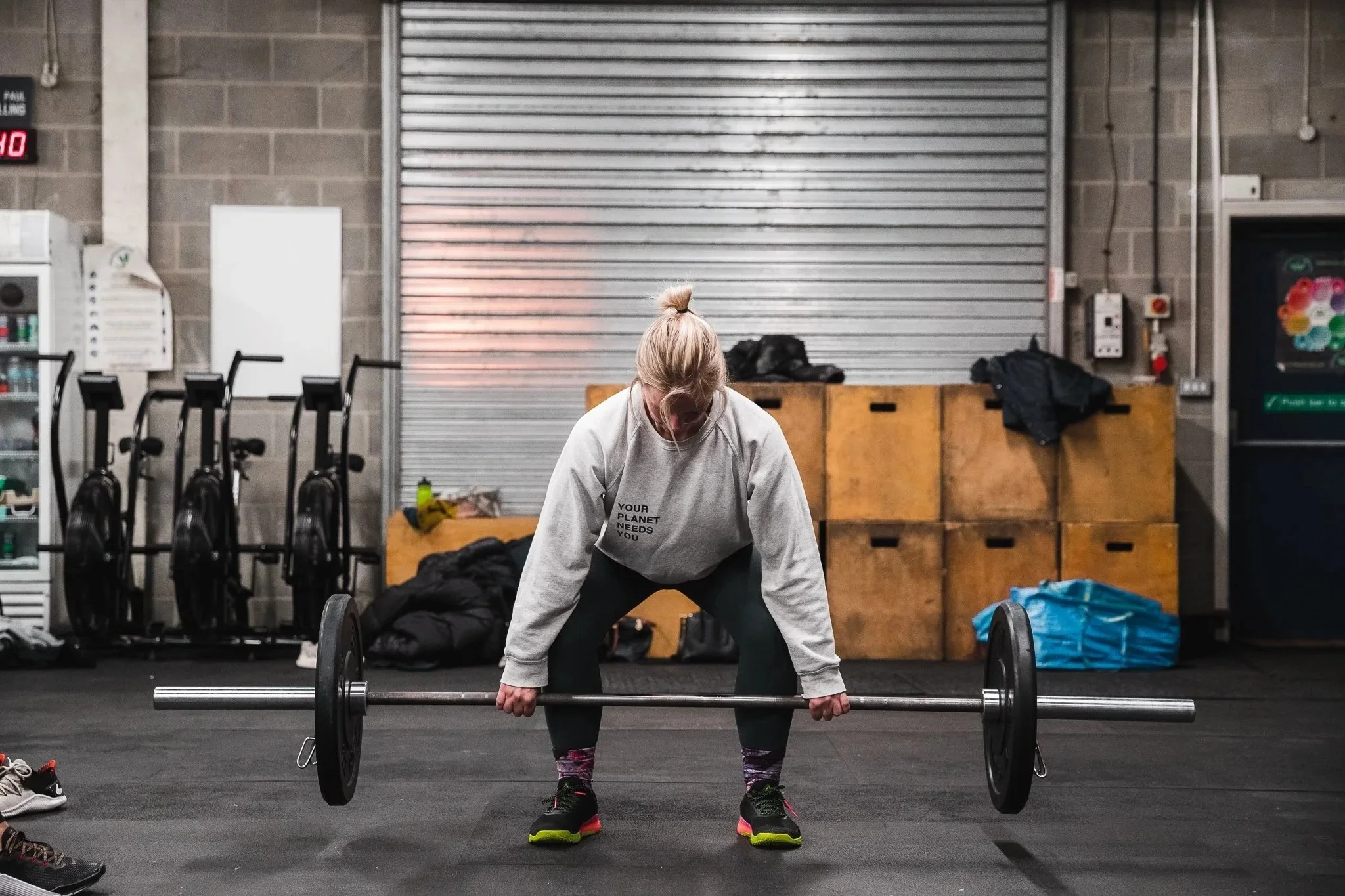 A woman with blonde hair tied in a bun lifting a barbell in a gym, standing on a black mat with gym equipment and a closed metal shutter in the background.