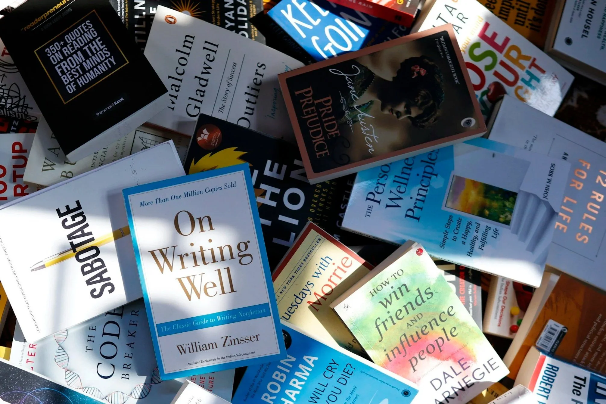 A pile of books on writing, self-help, and personal development, including titles like 'On Writing Well' by William Zinsser, 'The Code Breaker,' 'How to Win Friends and Influence People' by Dale Carnegie, and various other books about success, happiness, and psychology.