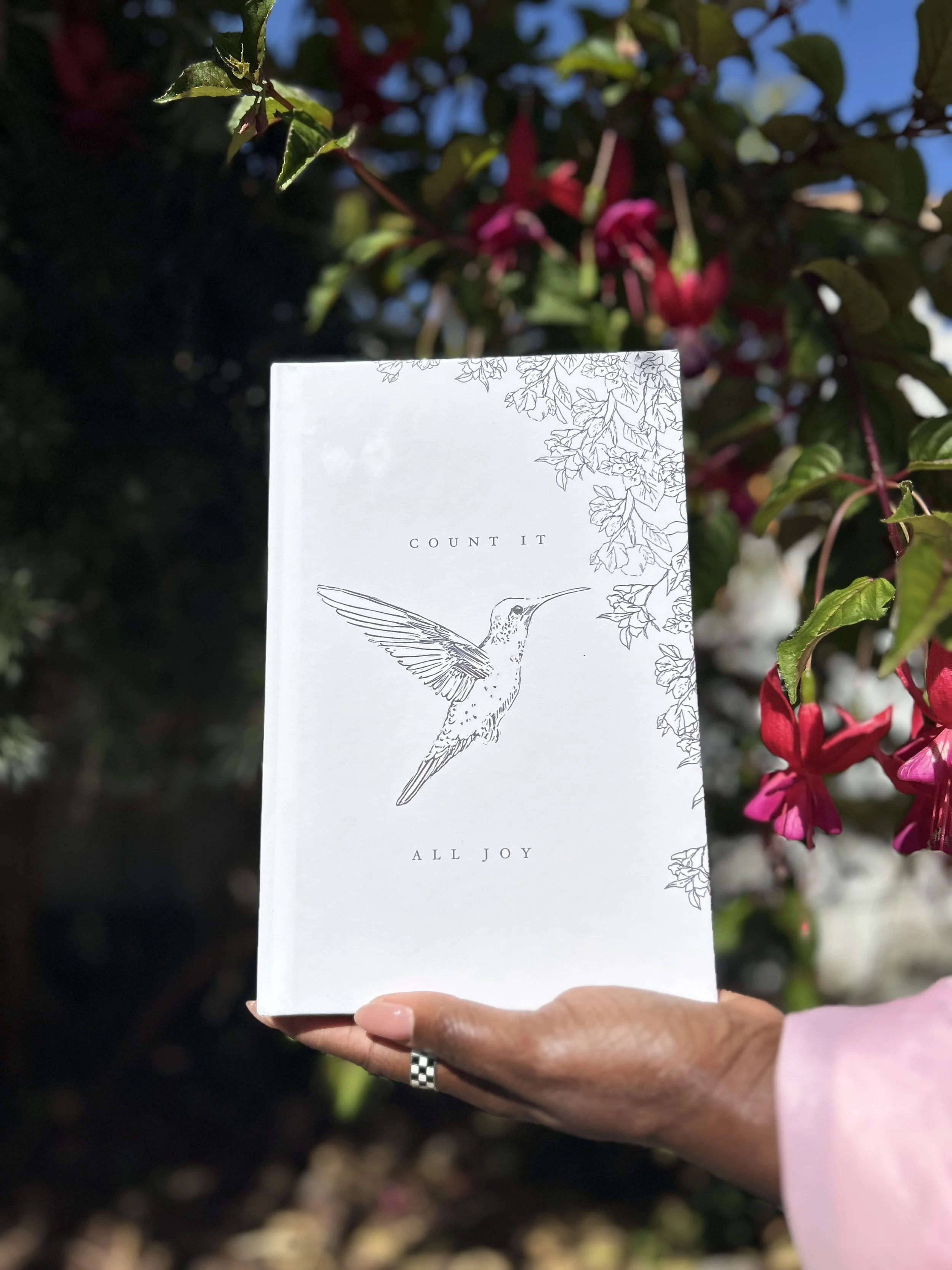 Hand holding a white book with a minimalist illustration of a hummingbird and floral details on the cover, with the phrases 'Count It' and 'All Joy'. The background features green leaves and pink fuchsia flowers under a blue sky.