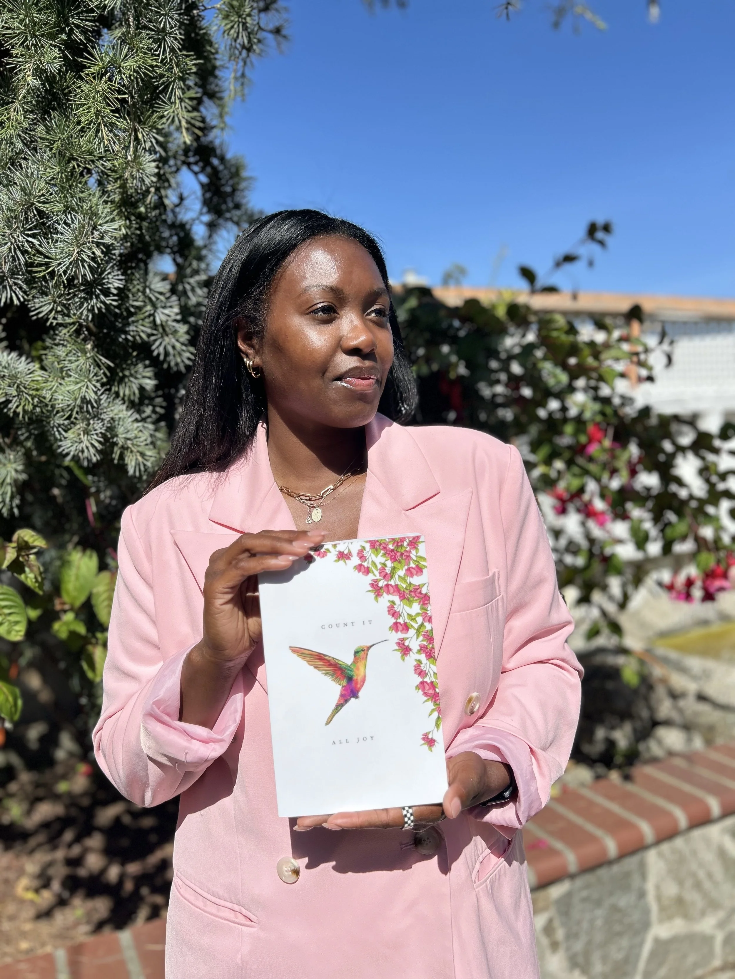 A woman with dark hair, wearing a pink blazer, holding a greeting card with a colorful hummingbird and pink flowers, standing outdoors with a tree, bushes, and a clear blue sky in the background.