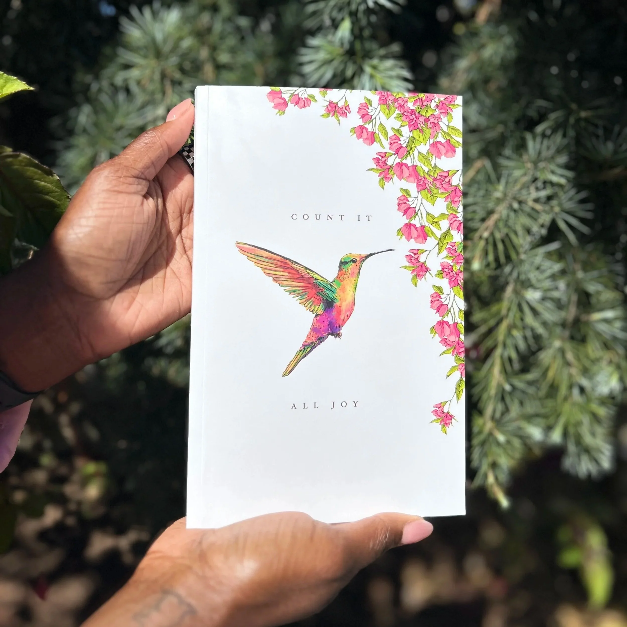 A hand holding a white notebook with a colorful illustration of a hummingbird and pink flowers, with the words 'Count It All Joy' on the cover, against a background of green foliage.