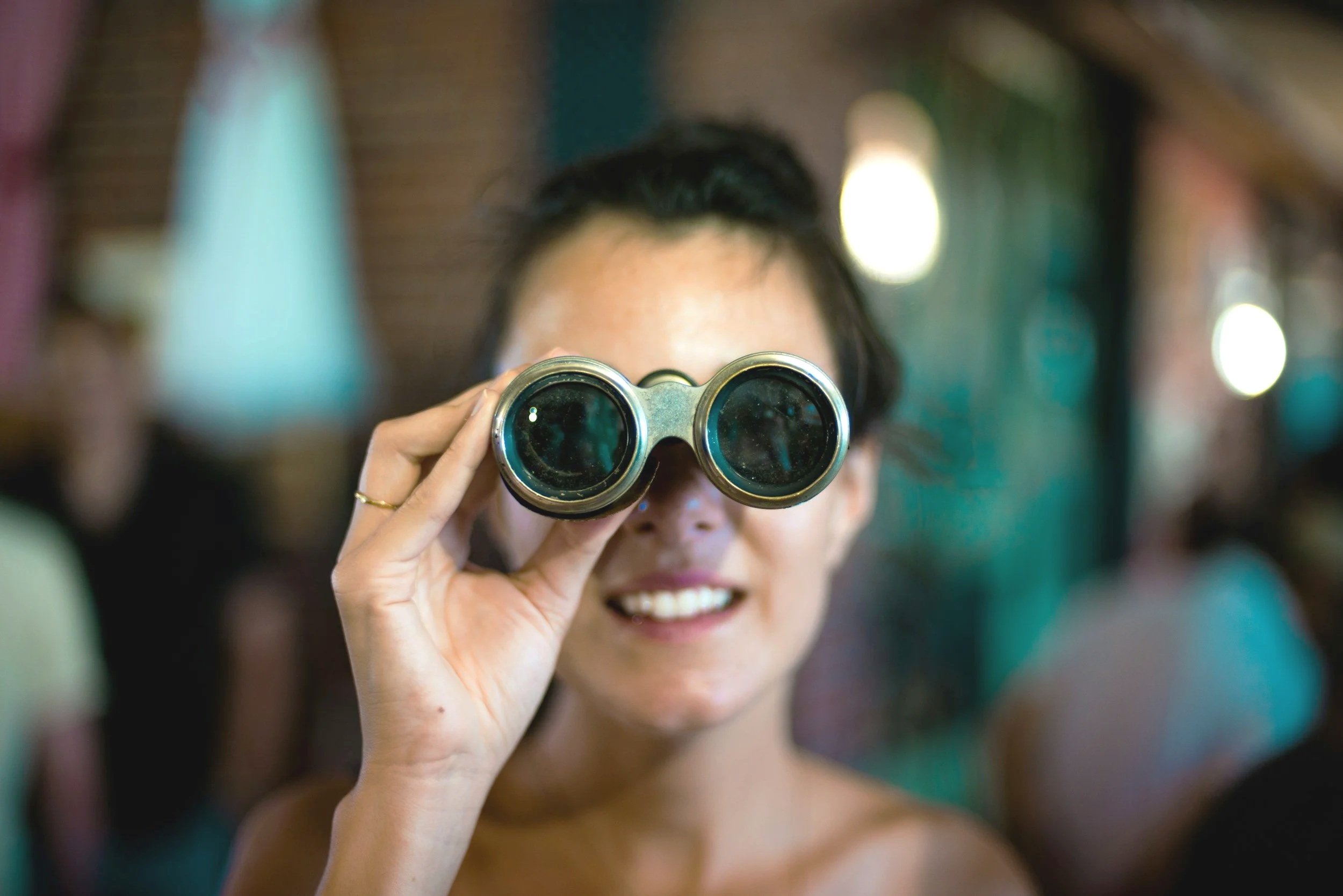 Woman smiling and looking through binoculars in a colorful indoor setting.