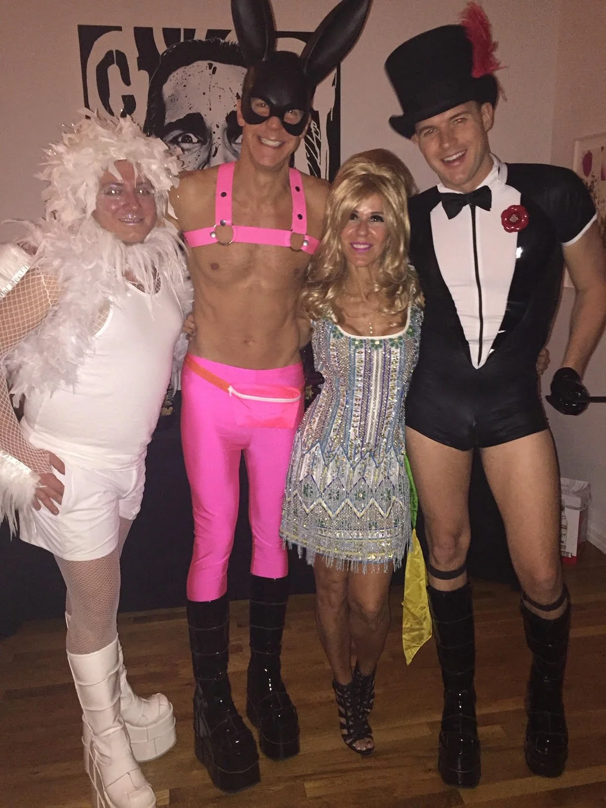 Group of five people dressed in colorful and elaborate costumes, standing indoors. One person wears a bunny costume with black ears and a mask, another is dressed in pink with platform boots, the third wears a sparkly dress, and the fourth is in a black tuxedo with a top hat and boots. They are smiling and posing for the camera.