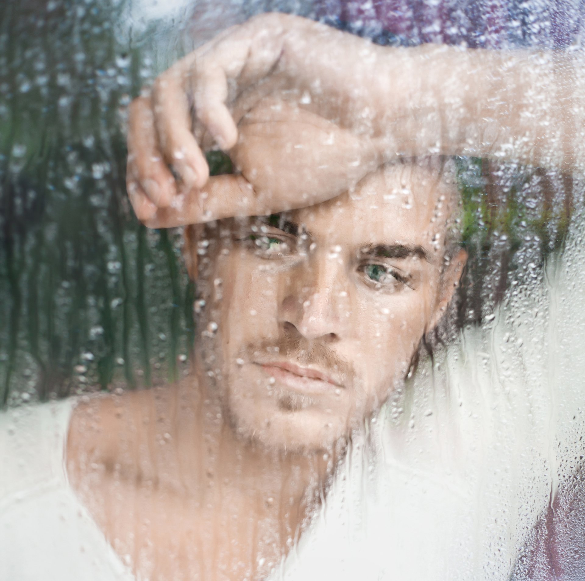 A young man with short hair and a slight beard looking through a rain-covered window, touching the glass with his hand.