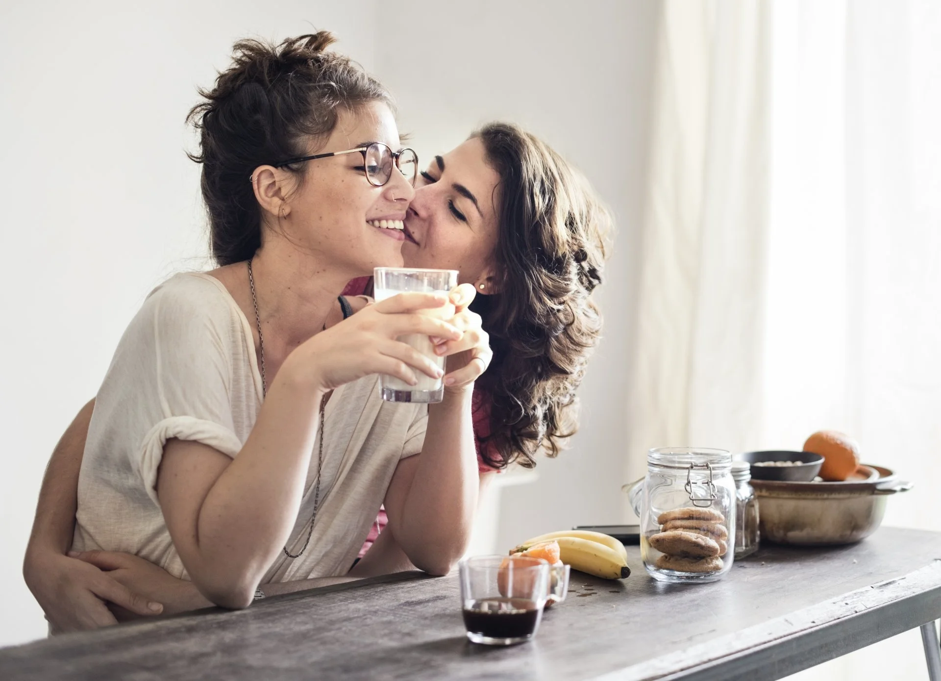 Two women sharing a joyful moment at a kitchen table, one drinking water and the other giving a kiss.