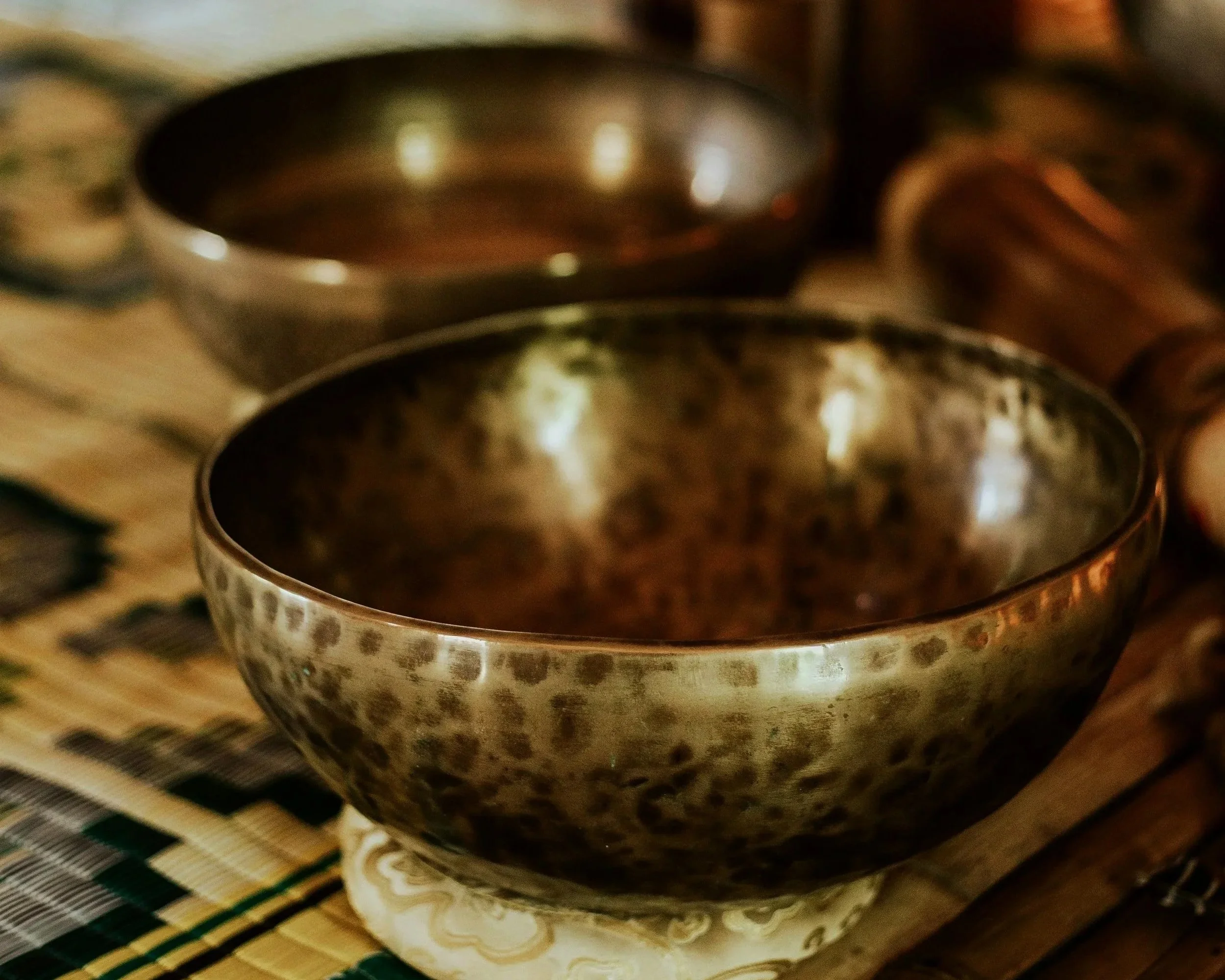 A close-up of a metallic singing bowl with a speckled pattern on a decorative base, with other similar bowls blurred in the background on a woven mat.