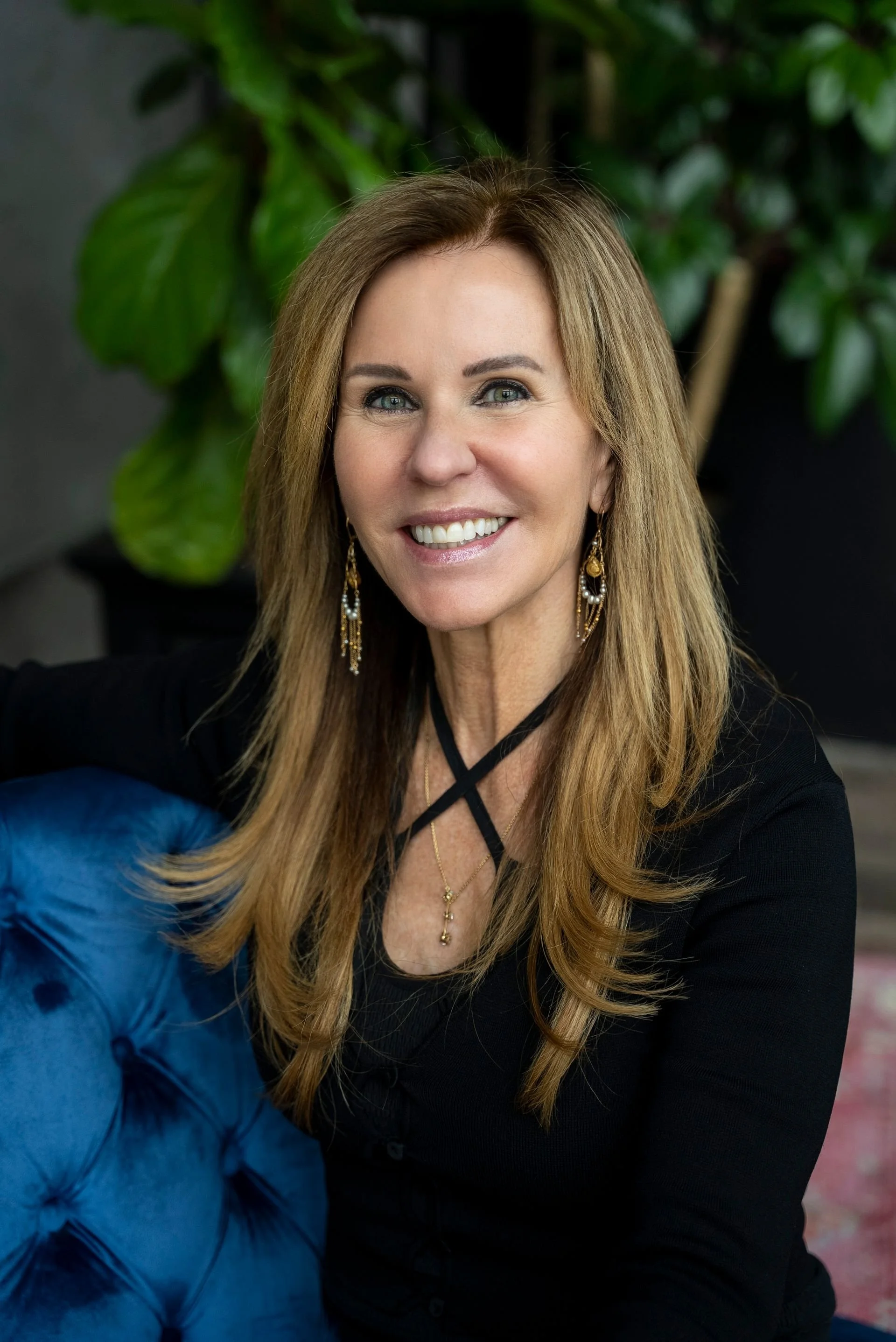 A smiling woman with long light brown hair, wearing a black top and statement jewelry, sitting next to a blue tufted chair with green plants in the background.