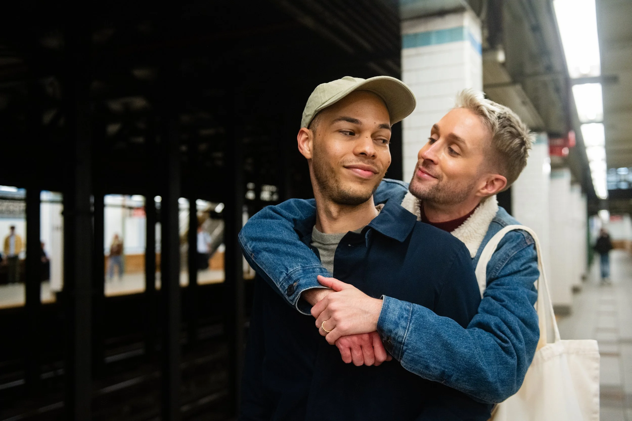 Two young married men embrace and smile at each other in a subway station.