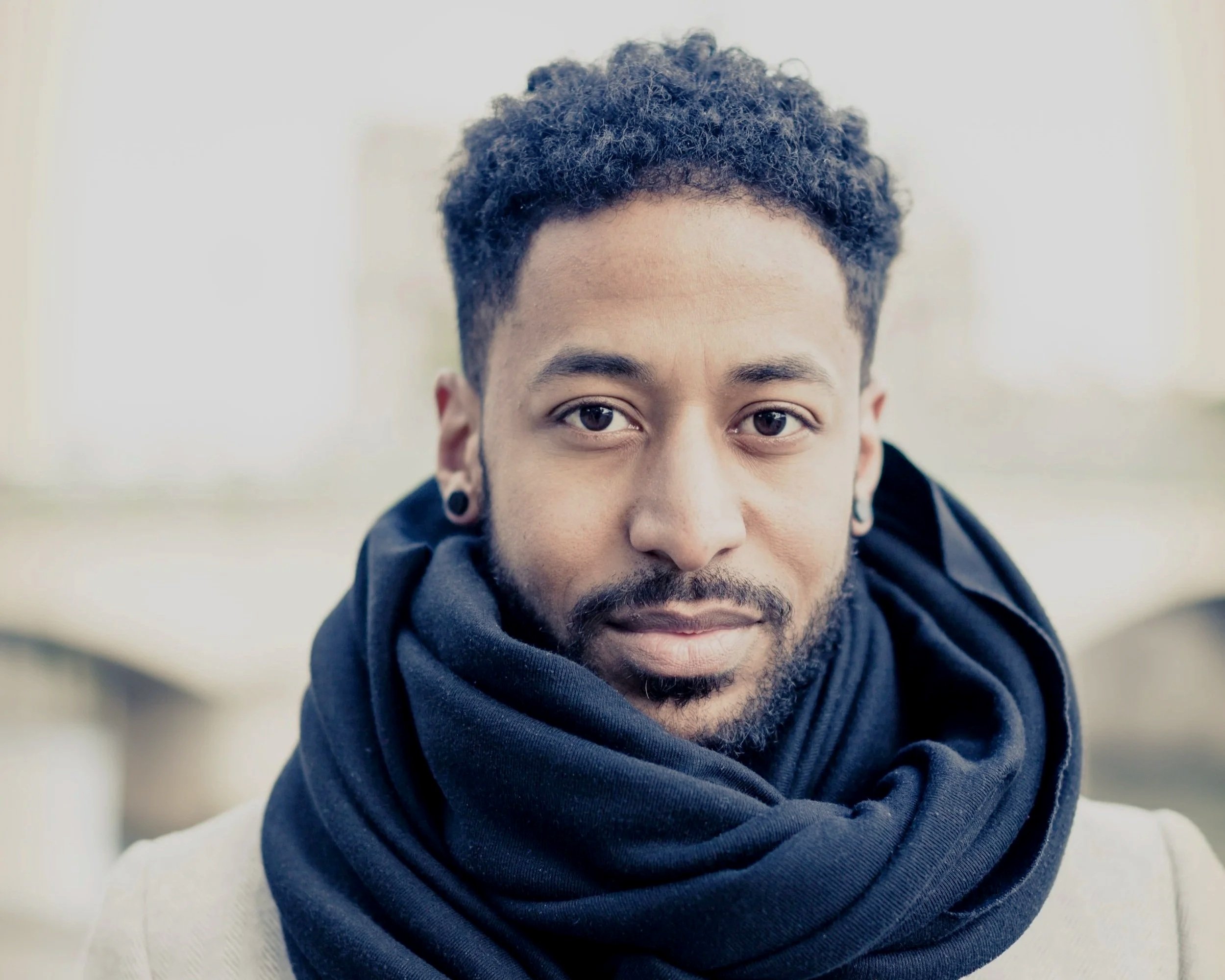 Close-up portrait of a young man with short curly hair, beard, and earrings, wearing a black scarf and beige jacket, outdoors with blurred cityscape background