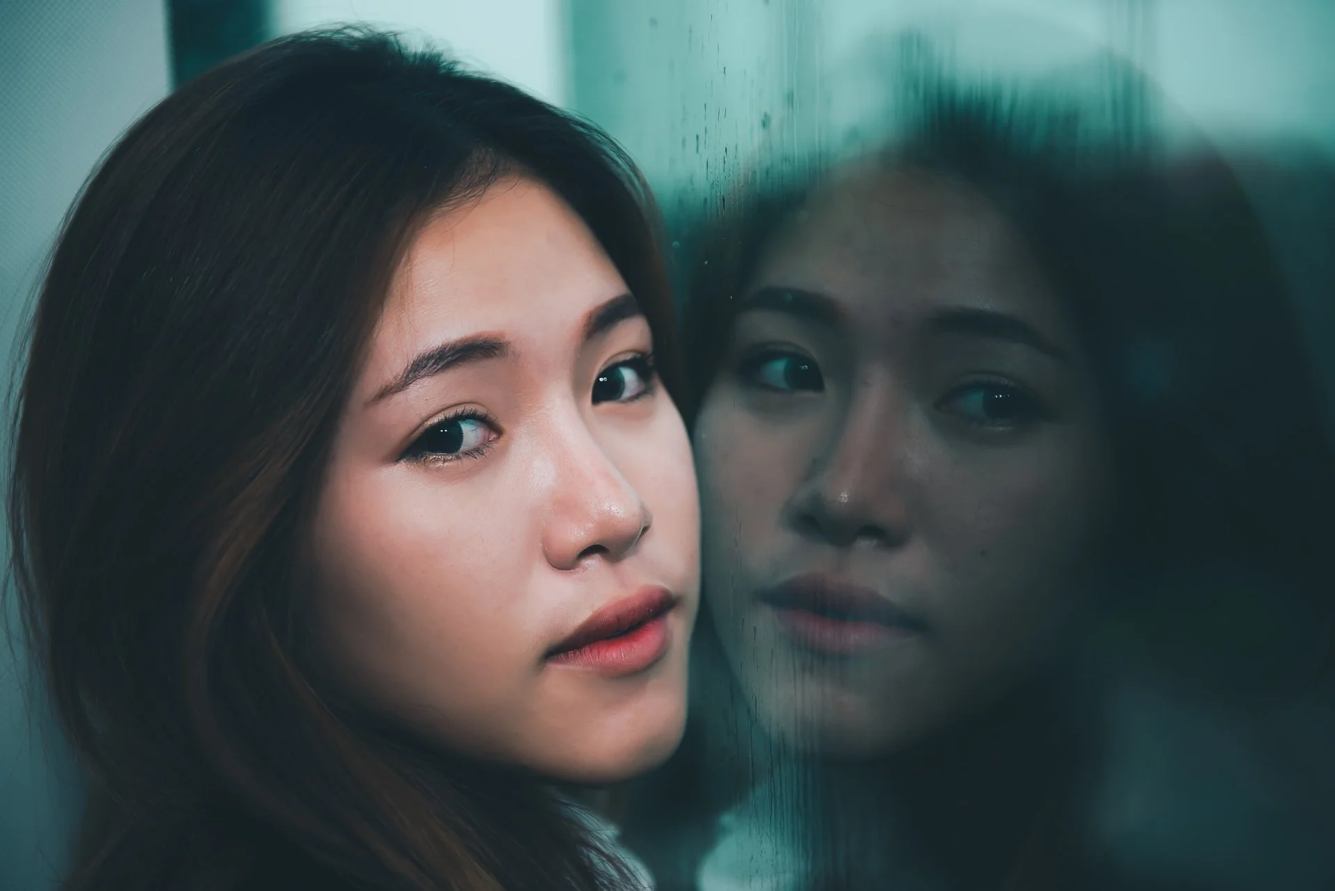 A young woman with long dark hair and light makeup looking at the camera near a reflective glass surface.