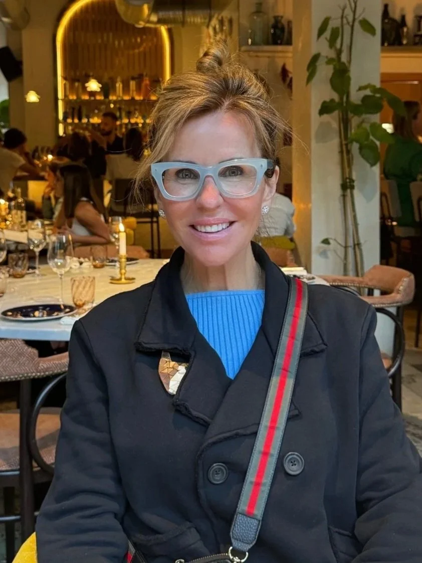 A smiling woman wearing clear glasses and a black coat sitting in a restaurant with a well-lit bar in the background.