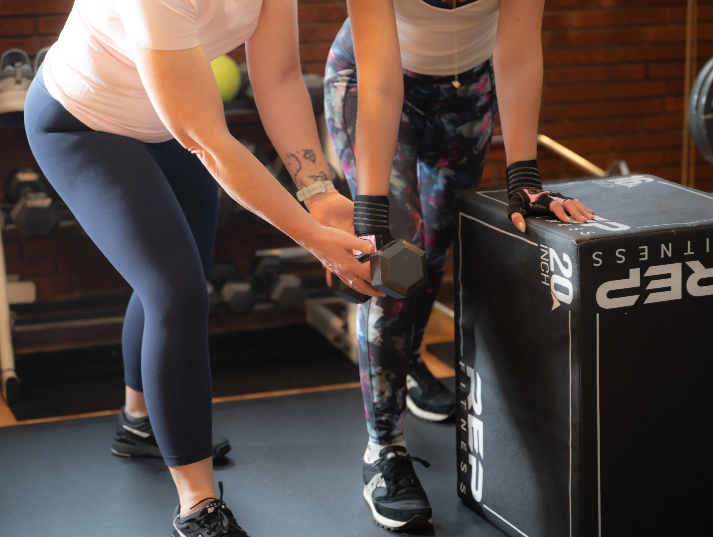 Two women high-fiving each other during a workout in a fitness studio with wooden floors and large windows.
