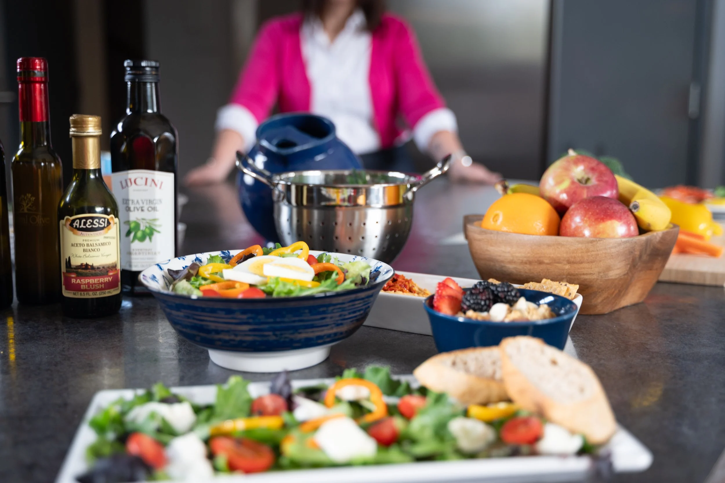 A breakfast spread on a white marble table includes bowls of oatmeal topped with fresh berries and flowers, waffles with toppings, a cup of tea, a glass of milk, a small jar of honey with a honey dipper, a small pot of red pepper flakes, and green foliage