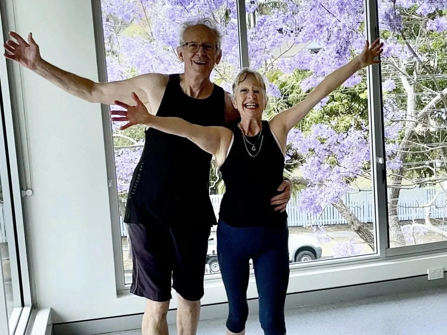 Two elderly women with athletic attire and sneakers, smiling and posing inside a room with large windows showing purple flowering trees outside.