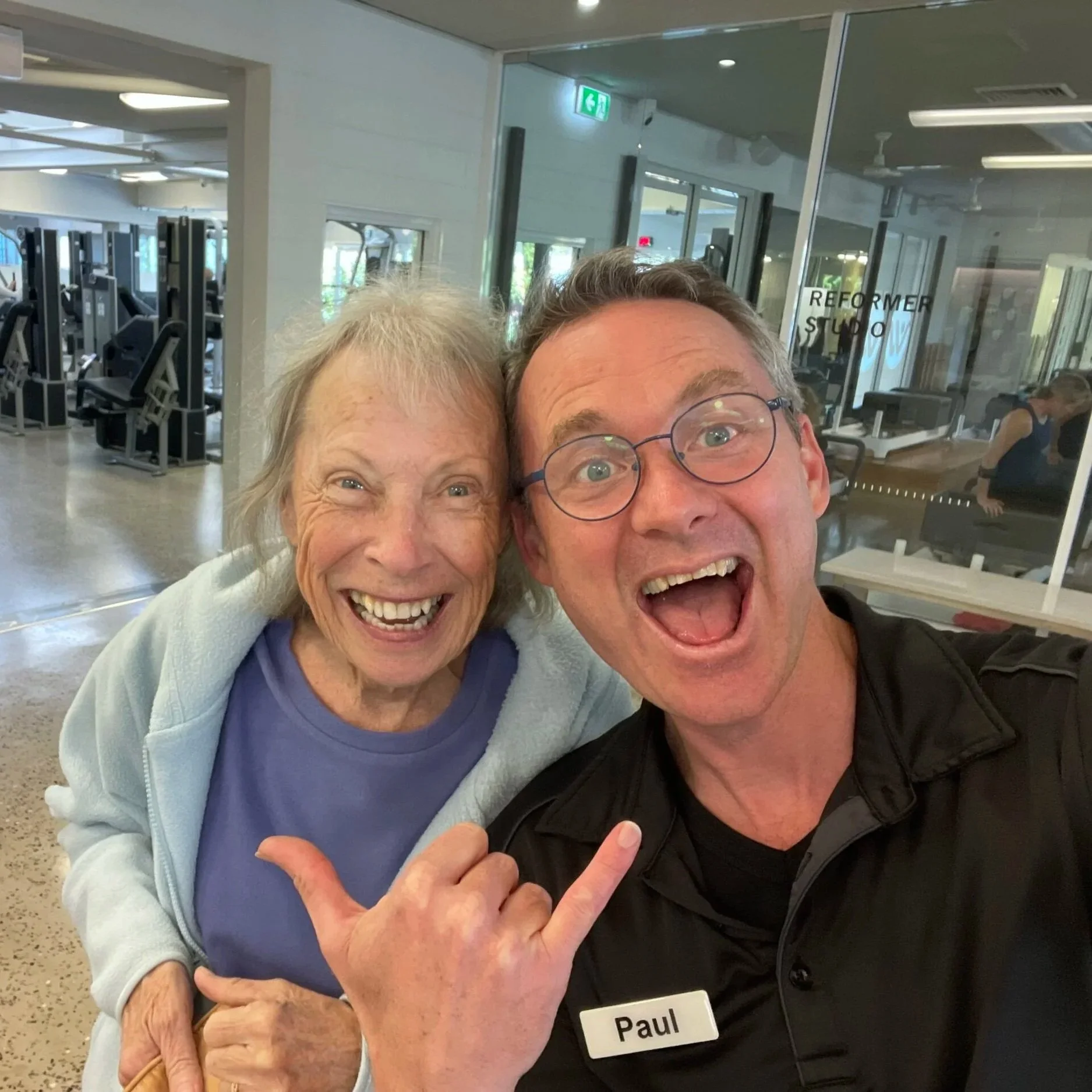A smiling elderly woman and a man wearing glasses, with a name tag that says Paul, taking a selfie together in a gym or fitness center.