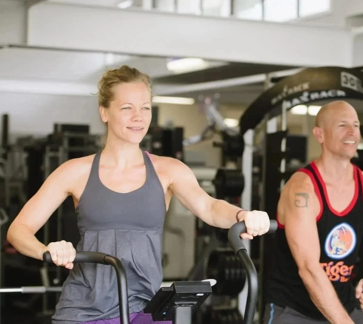 A group of people participating in a fitness class, smiling and looking at the camera, in a gym setting.
