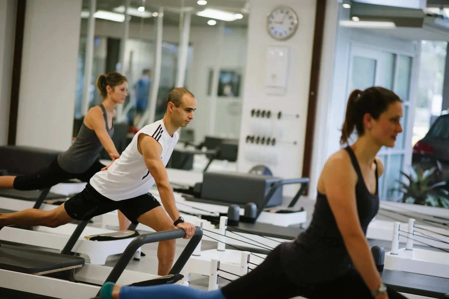 A woman exercising on a Pilates reformer machine in a bright, spacious room with large windows showing outdoor greenery.